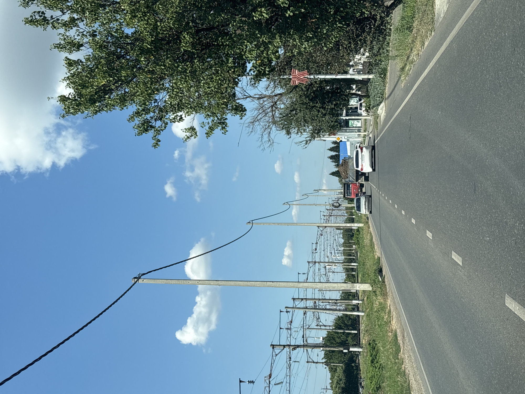 A sunny day is depicted with a road leading towards a horizon dotted with trees and buildings. Power lines crisscross the sky, supported by concrete poles. Several vehicles, including cars and a truck carrying soil, are visible on the road. A large tree with lush green leaves stands prominently on the right side of the frame, while a red ribbon-like decoration hangs from a pole in front of it. The road is asphalted and divided by dashed white lines. The environment appears to be a suburban or semi-rural area, possibly on the outskirts of Yuzhny, Russia, given the presence of such infrastructure.