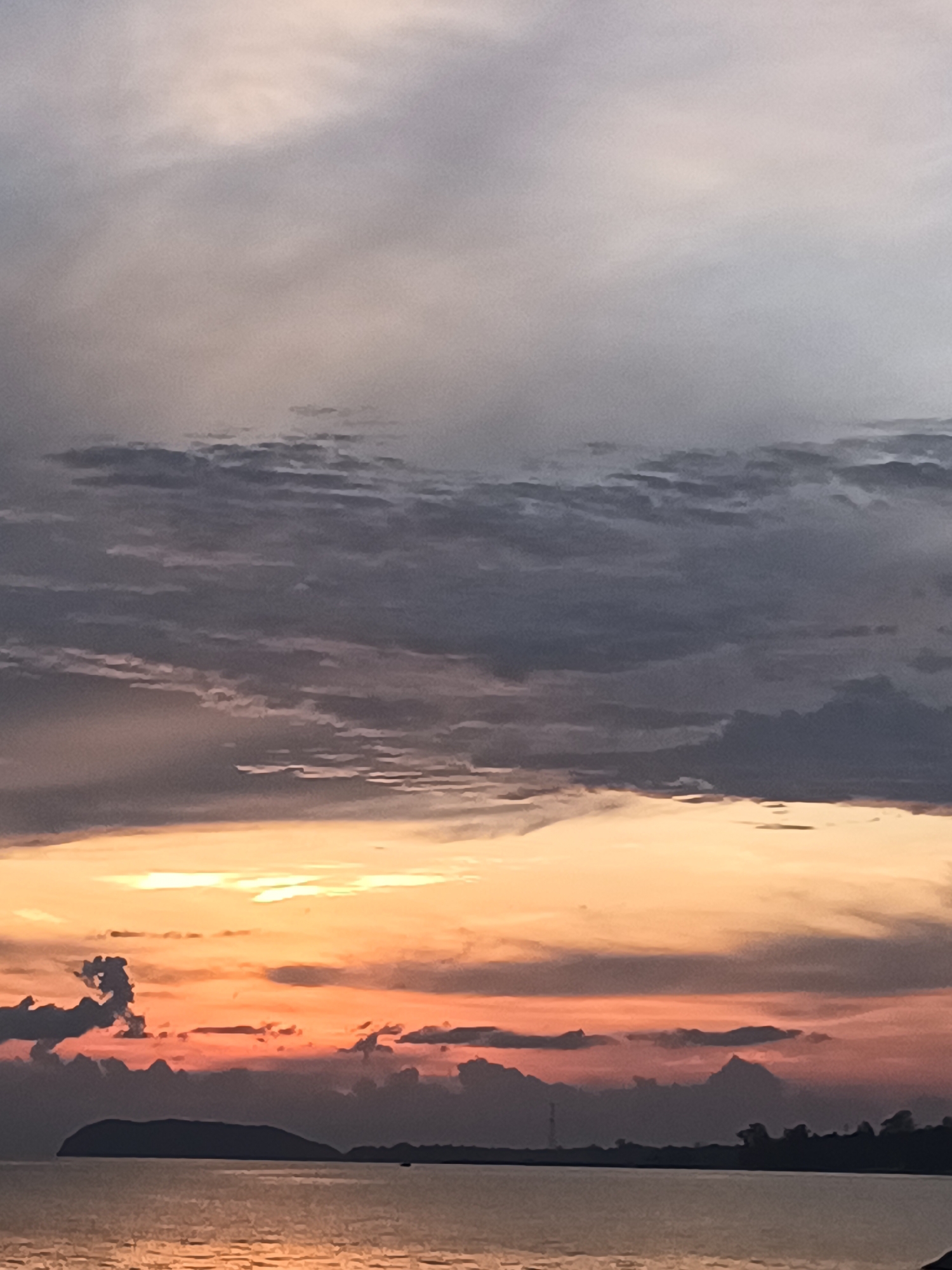 The image shows a sunset over the ocean in Baan Kai, Thailand. The sky is filled with clouds, colored in shades of gray, orange, and red. The sun is setting, casting a warm glow over the scene. In the distance, there is a dark silhouette of an island and trees on the shoreline. The water is calm and reflects the colors of the sky.