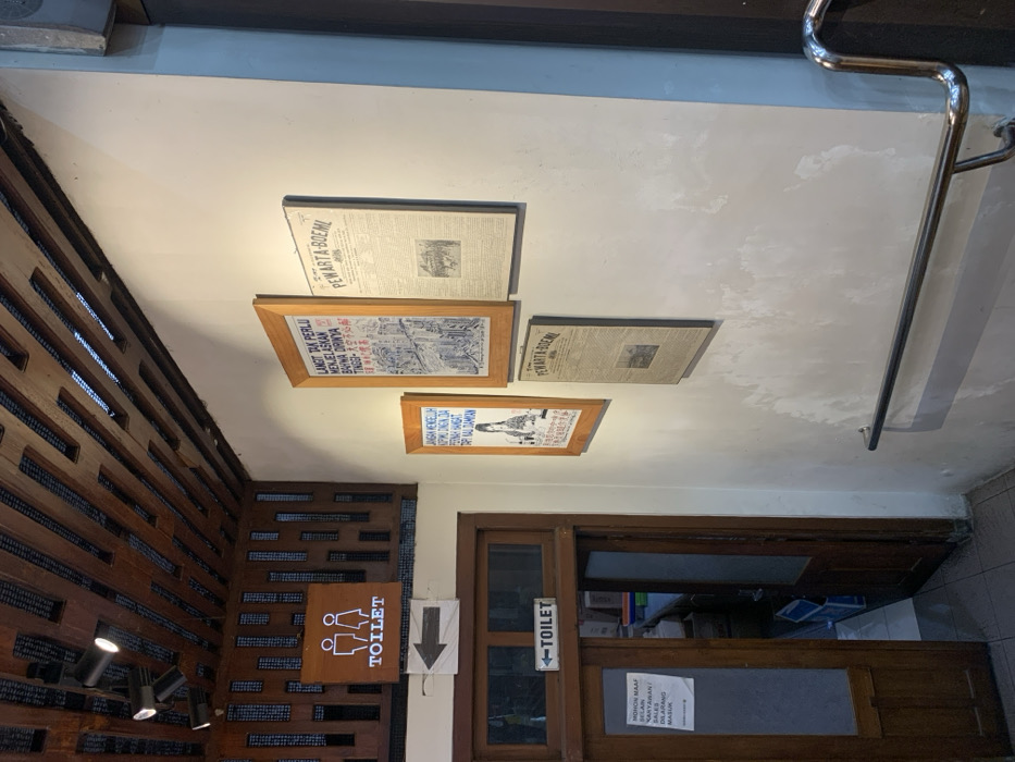 The image shows the interior of a building in Muntilan, Indonesia, located at latitude -7.584 and longitude 110.287. Three framed posters, two of which are titled "PEWARTA BOEMI," are hung on a white wall. There is a wooden door with a "TOILET" sign above it. An arrow points toward the door, and a sign on the door reads "MOHON MAAF SELAIN KARYAWAN PT. KAI DILARANG MASUK." A stainless steel handrail runs along the wall to the right of the door. A wooden, slatted ceiling is visible above. The environment is a dimly lit, indoor hallway.