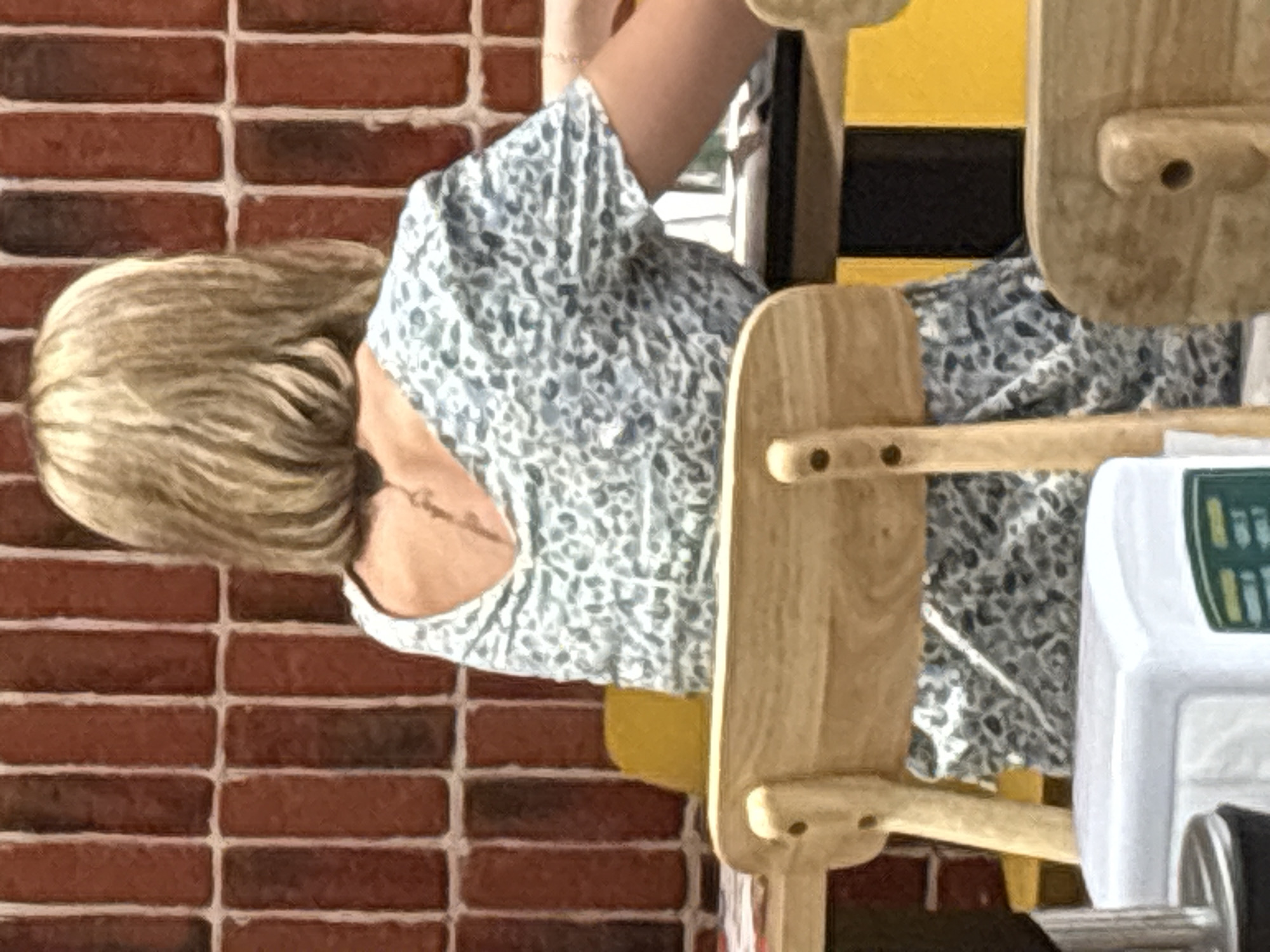 The image shows a woman with short, blonde hair seen from behind, wearing a light-colored, patterned dress. She is seated at a table in what appears to be a restaurant or cafe. A tattoo is visible on her upper back. The background consists of a red brick wall. In the foreground, a wooden chair back and a white object, possibly a napkin dispenser, are visible. The environment suggests an indoor setting. There is no visible information in the image to determine a specific location in the city of Astana, Kazakhstan.