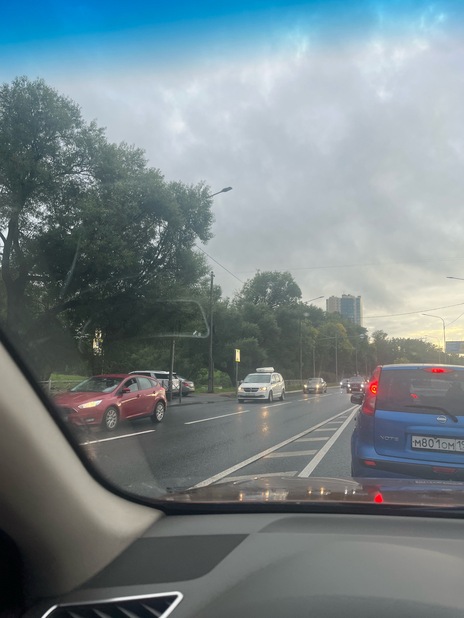 The image captures a street scene in Saint Petersburg, Russia, likely taken from the interior of a vehicle. The sky is overcast with grey clouds, suggesting a diffused or possibly rainy day. The road is wet, reflecting the headlights of the cars and streetlights, indicating recent precipitation.
Several vehicles are present on a multi-lane road. In the foreground, from the perspective of the camera's vehicle, a blue Nissan Note is visible in the right lane, with its license plate reading "M801OM 19 RUS". Further ahead, a line of traffic is moving forward. Cars observed include a white minivan in the left lane, a silver car, and a red hatchback in the far left lane, which appears to be a Ford Focus.
Trees line the left side of the road, with dense foliage suggesting a park or a tree-lined boulevard. In the distance, several modern high-rise buildings are visible, peeking above the trees, indicating an urban setting. Streetlights are positioned along the road. The overall impression is one of a typical urban commute on a cloudy day.