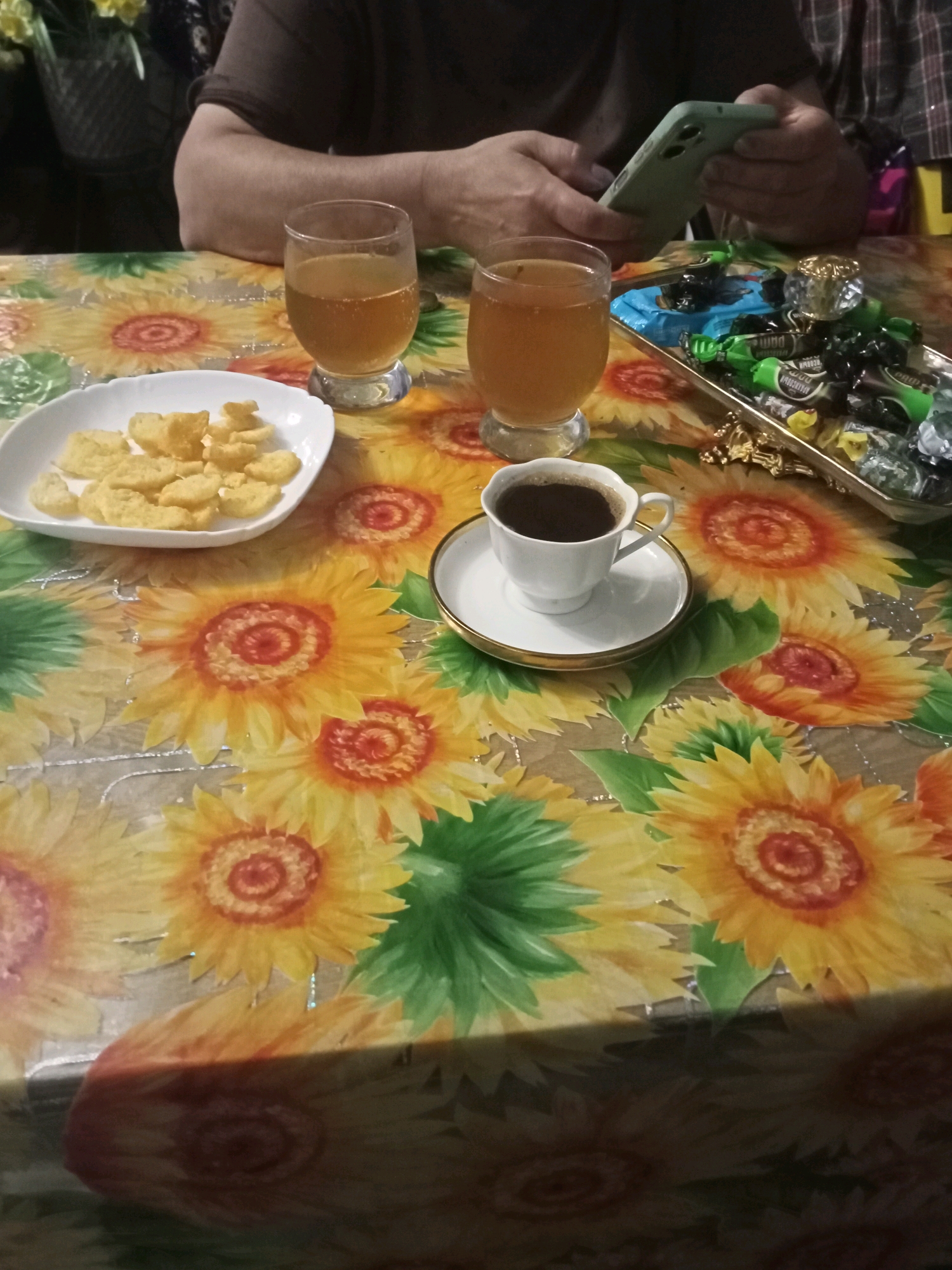 A close-up shot shows a person's hands holding a smartphone and resting on a table covered with a sunflower-patterned tablecloth. On the table, there are two glasses of amber liquid, a white bowl filled with light-colored snacks, and a white cup of dark liquid with a saucer. To the right, a rectangular tray contains an assortment of wrapped candies in green and black wrappers, topped with a crystal-like object. The scene appears to be set indoors, with the background out of focus, hinting at a domestic setting in Tsandrypsh, Abkhazia.