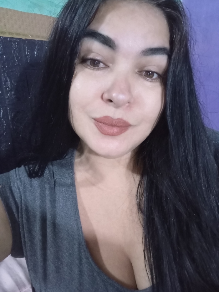 A woman with long, dark hair frames her face. She has light skin, brown eyes, and wears a muted brown lipstick. She is wearing a gray tank top. The background includes a mottled black and silver fabric and a purple wall. The lighting is soft, highlighting her features. The overall impression is casual and intimate, capturing a close-up portrait.