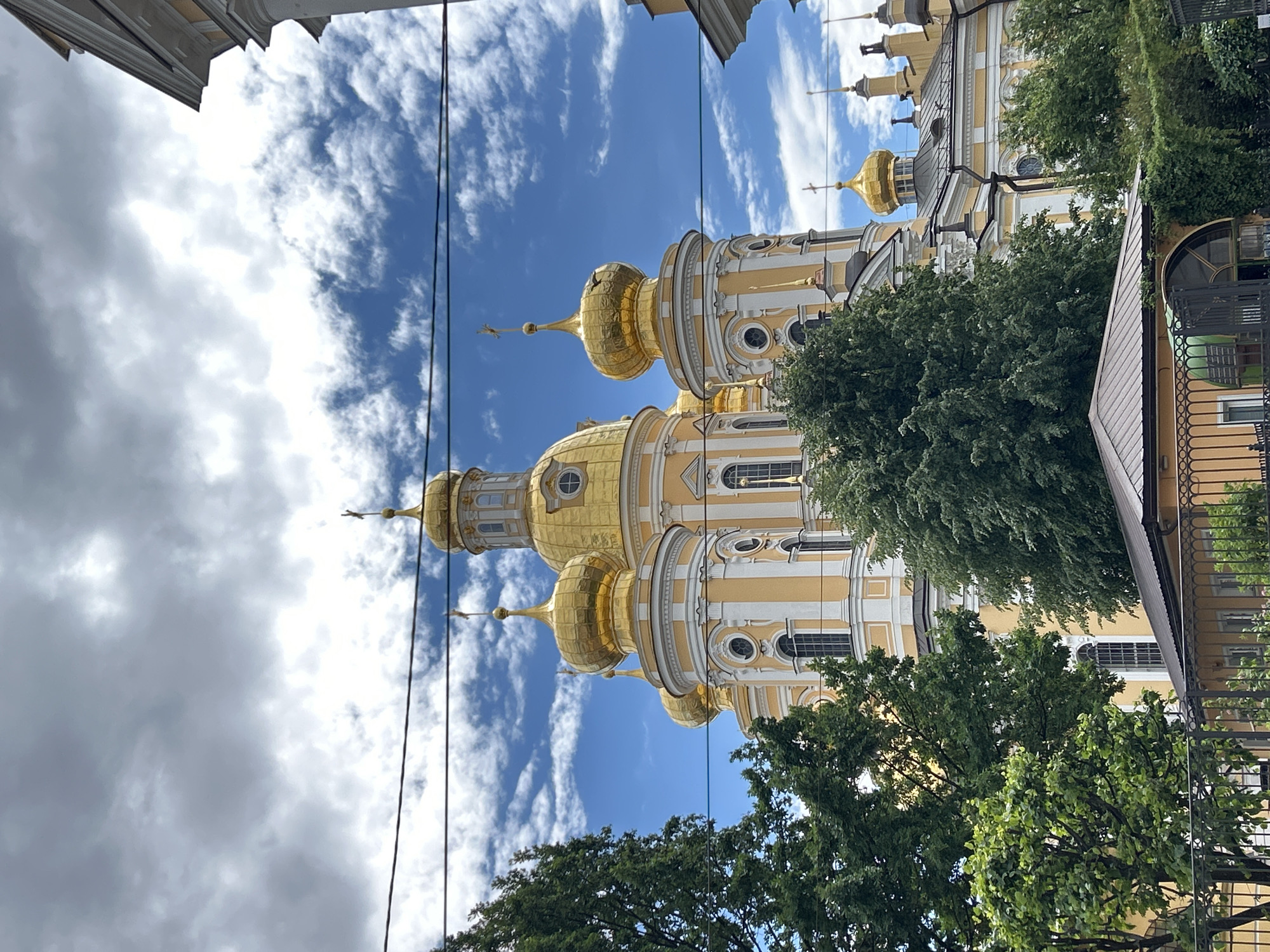 The image captures a low-angle view of a grand, ornate building with multiple golden domes, set against a bright blue sky dotted with white, wispy clouds. The architecture appears to be of a religious or historical nature, possibly a church or cathedral, characterized by its yellow and white facade, arched windows, and intricate decorative elements. Several trees with lush green foliage partially obscure the lower portions of the building, suggesting an outdoor setting in a potentially urban or park-like environment. Power lines are visible in the foreground, cutting across the scene and adding a modern element that contrasts with the historical architecture. The lighting suggests it is daytime, likely with bright sunlight illuminating the golden domes and the sky. The overall impression is one of architectural splendor and a serene, possibly historical, setting. No people are visible in the frame, and no text is legible.
