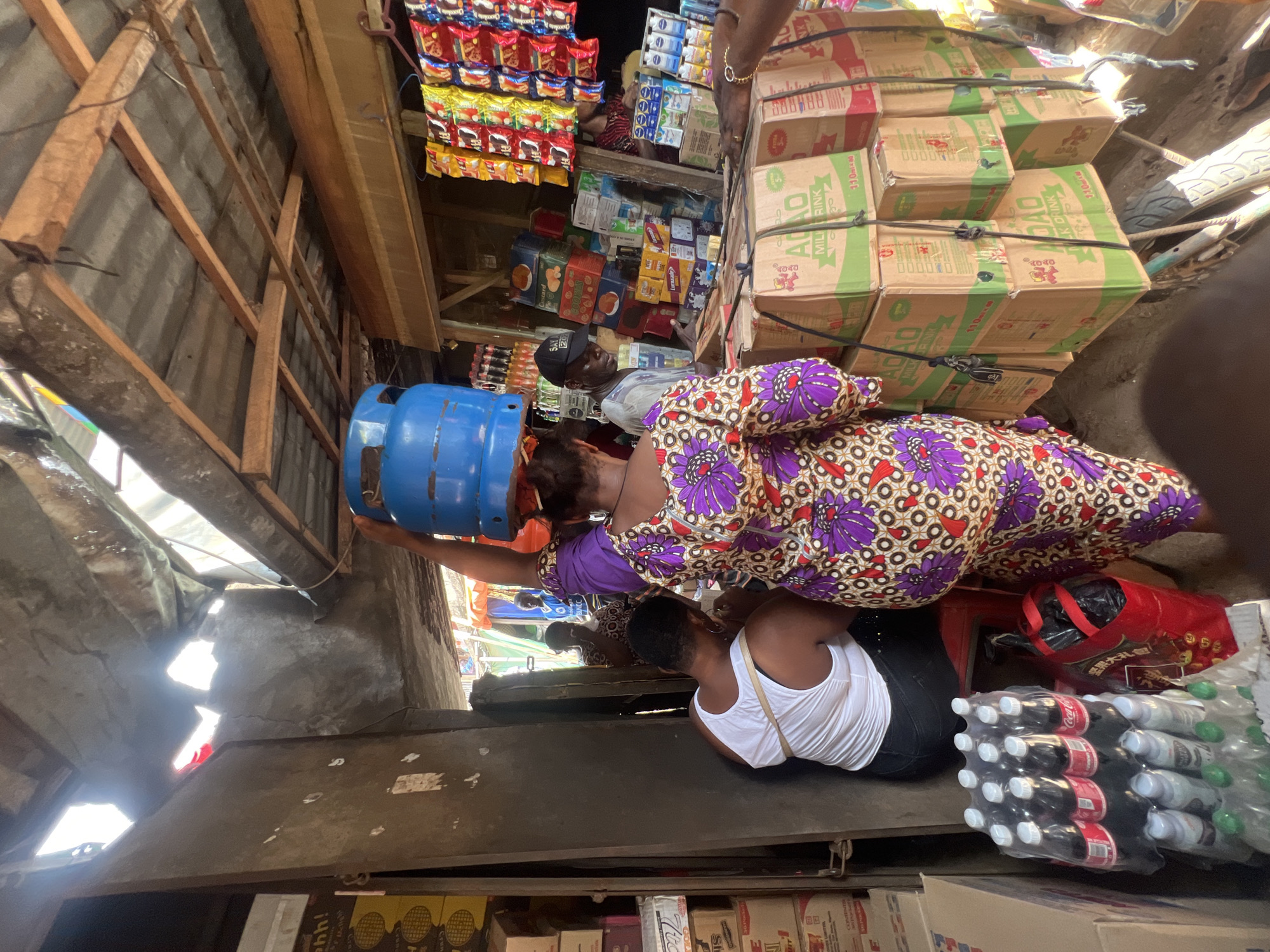 The image depicts a bustling scene inside a small shop or market in Nkpor, Nigeria. A woman wearing a colorful purple and brown patterned dress is carrying a blue gas cylinder on her head. Another woman is sitting on a red plastic stool near stacks of bottled drinks (Coca-Cola and others). The shop is packed with various goods, including boxes of "ADADO MILK RINK" stacked high, snacks hanging on display, and other packaged items. The shop appears to be situated under a makeshift roof, constructed from wooden frames and corrugated metal sheets. The lighting is dim and uneven, suggesting an indoor setting with limited natural light. The environment is crowded, indicating a busy marketplace.