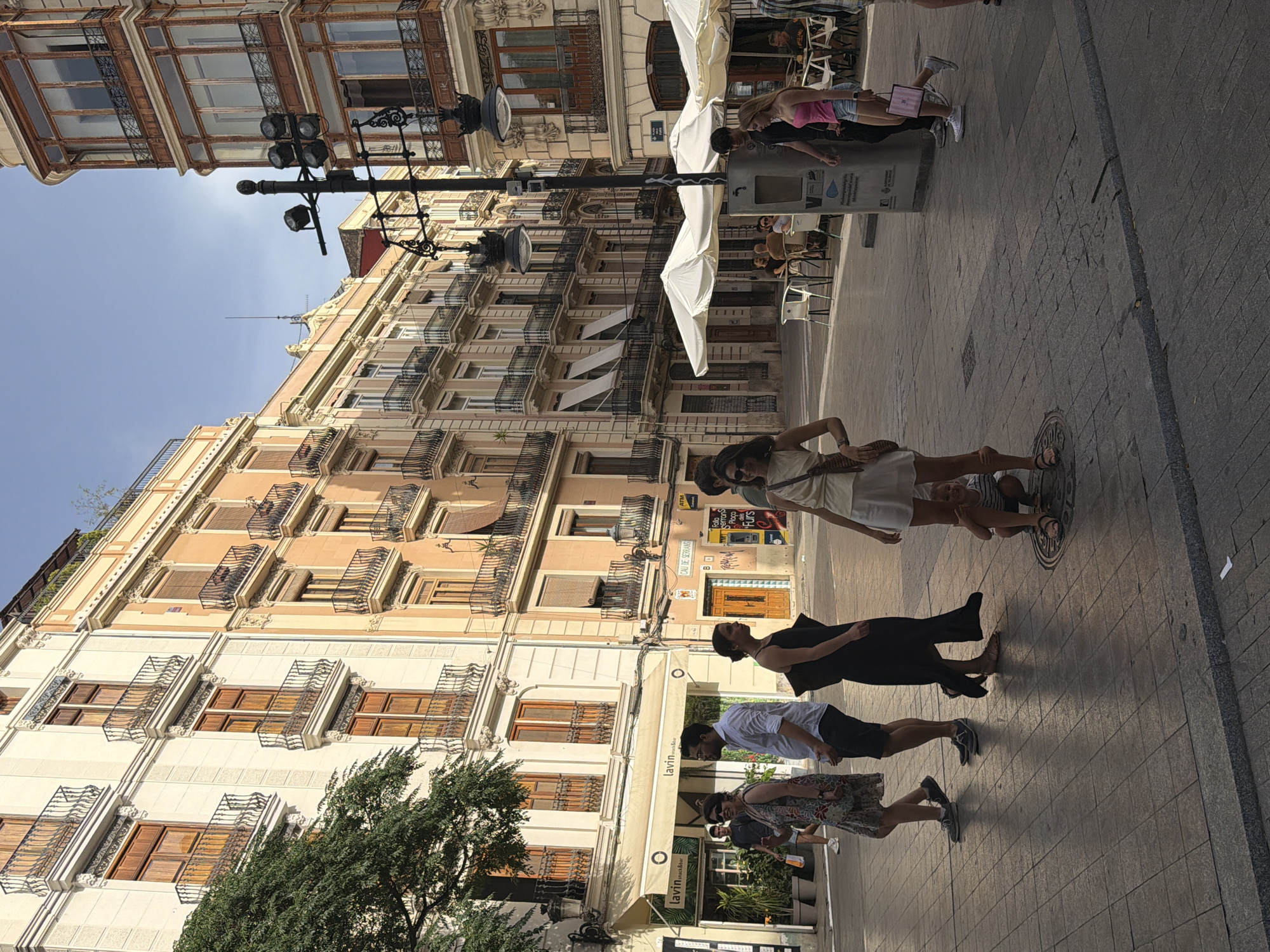 In a city square, people walk past buildings with balconies and awnings. In the foreground, a woman in a white dress walks toward the viewer. To her left, a man in a white shirt and dark pants walks away. A woman in a patterned dress walks to his left. A child is crouching behind the woman in the white dress. To the right, a couple walks past a trash can. In the background, people sit at tables under umbrellas. The buildings are tall and ornate, with many windows. The sky is clear and blue. The ground is paved with square tiles.