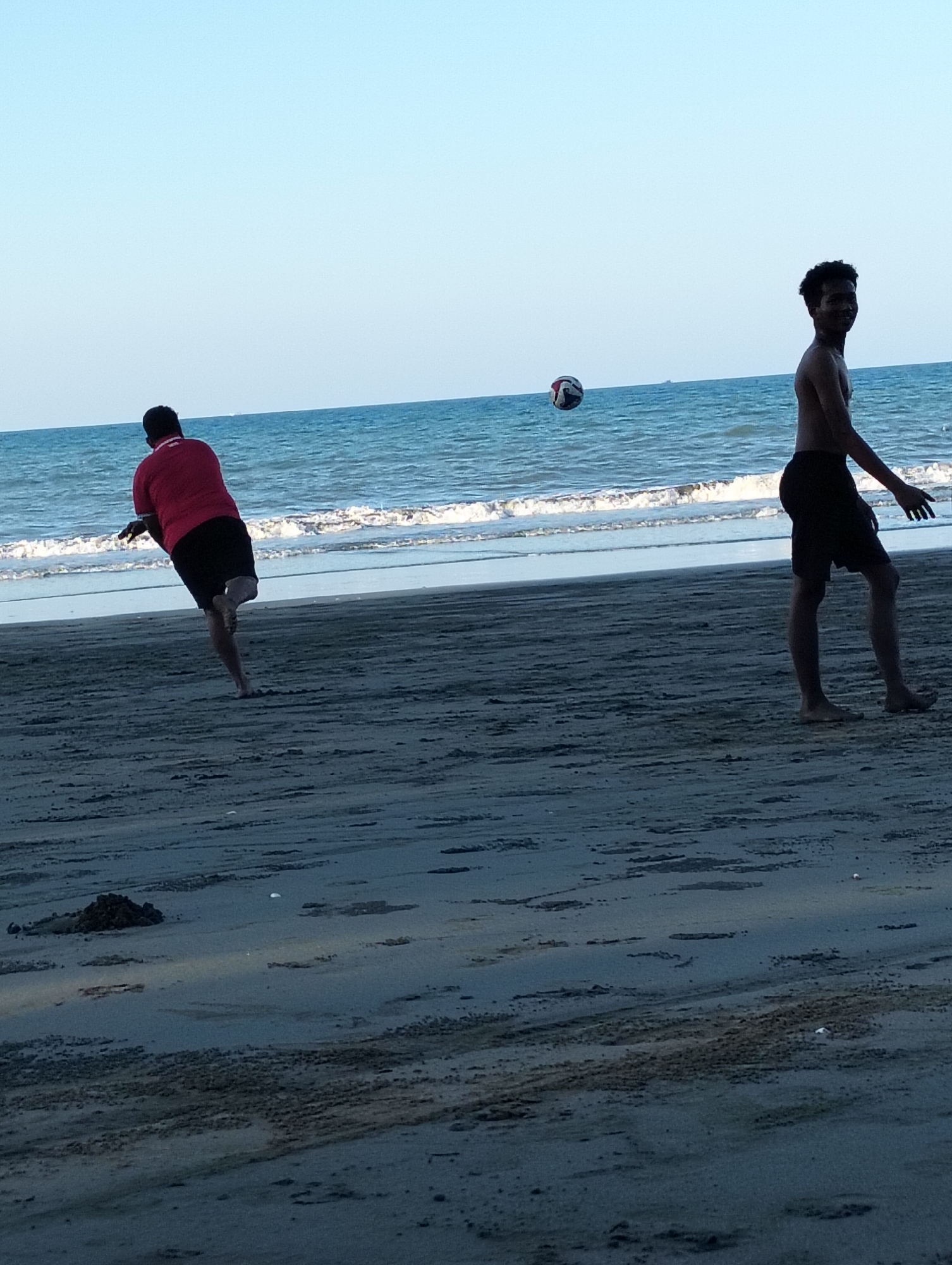Two young men are playing with a volleyball on a sandy beach in front of the ocean under a clear sky. The man on the left, wearing a red shirt and black shorts, is in mid-action, with his back to the camera, about to hit the ball, which is in the air. The man on the right, shirtless and wearing black shorts, is looking towards the camera with a slight smile. The ocean is visible in the background with gentle waves. The beach sand appears dark and wet in some areas, indicating it might be close to the water line. There are some small objects, possibly debris or shells, scattered on the sand. The location is identified as Peukan Sot, Indonesia.