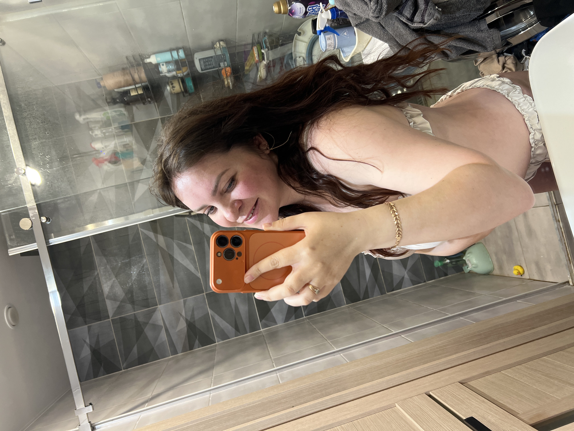 A woman is taking a selfie in a bathroom. She is holding an orange smartphone with three camera lenses in her right hand. She has brown hair and is wearing a white top and a gold bracelet on her right wrist. The bathroom features grey patterned tiles on the walls and beige wood-paneled cabinets. A glass shower door is visible on the left side of the image. Bathroom supplies are visible in the background. The location in the city of Noviy, Russia, cannot be determined from the given image.