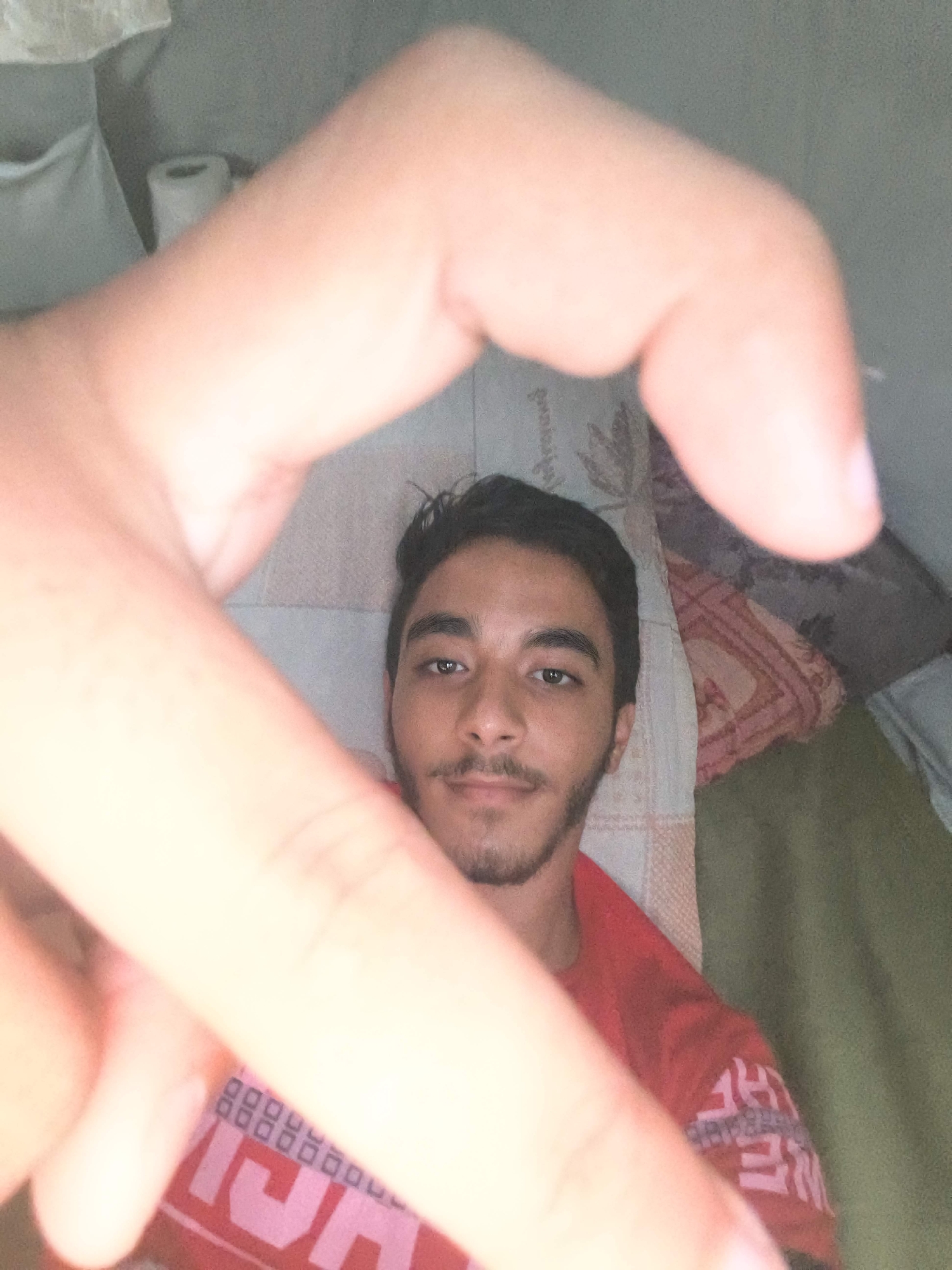 A close-up selfie shows a young man with dark hair and a beard, wearing a red t-shirt. His left hand is held up, obscuring part of his face and creating a heart shape with his fingers. The background is blurry, but appears to be a room with a wall, a roll of toilet paper, and some patterned fabric. The man is looking directly at the camera with a neutral expression. The image is lit by an overhead light source, casting shadows on his face.