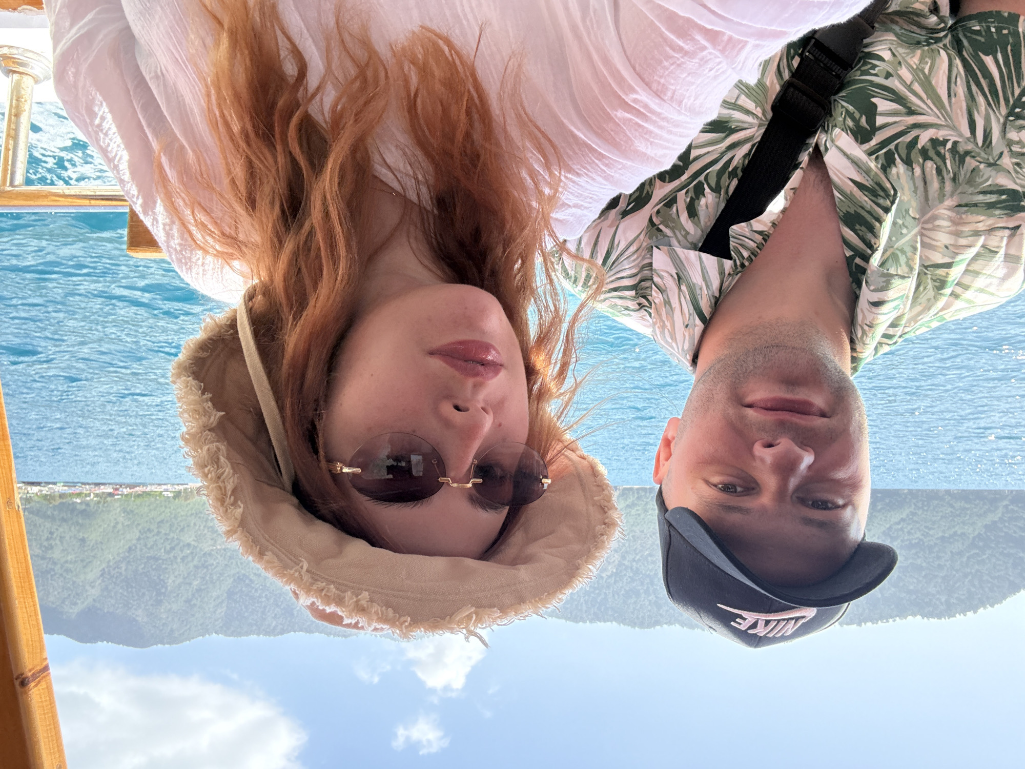 A couple is taking a selfie on a boat. The woman has long, red hair and is wearing a white shirt, a beige hat with a fringed brim, and sunglasses. The man is wearing a green and white leaf-patterned shirt and a black "Nike" baseball cap with a pink logo. They are positioned close together, facing the camera. The background shows blue water, a mountainous coastline, and a clear sky with some clouds. The location appears to be near Kumluca, Turkey, based on the landscape visible in the background.