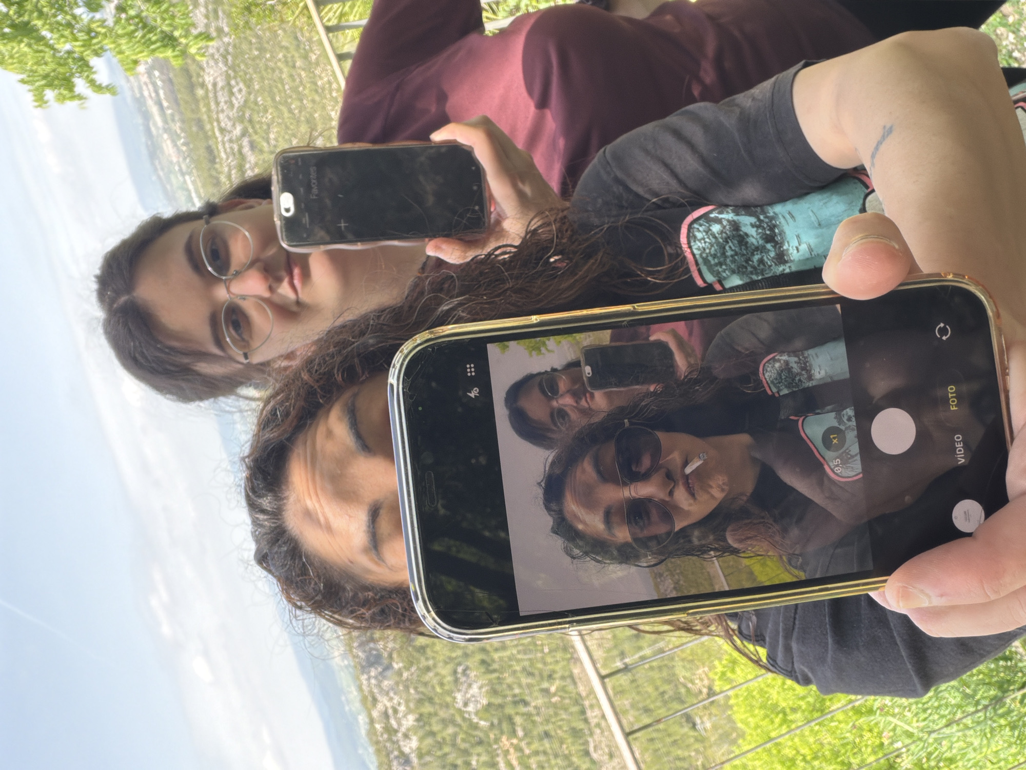 The image, oriented sideways, captures two individuals, likely young women, outdoors at a scenic overlook in Olèrdola, Spain.
The primary subject in the foreground is holding a smartphone, capturing a reflection. The phone's screen prominently displays a "selfieception," showing the foreground woman and a second person reflected, within a camera interface. The foreground woman has dark, curly hair, wears sunglasses, and has a lit cigarette in her mouth. She is wearing a dark t-shirt with a light blue and green graphic design. A small, unreadable tattoo is visible on her right forearm.
The second individual, partially visible behind the foreground woman and also reflected in the phone screen, has dark, straight hair and wears round-rimmed glasses. She appears to be holding a second smartphone, which is partially visible. This phone's screen displays "Favorites" and a "+" icon, suggesting an app menu.
The activity depicted is taking a reflection selfie, creating a recursive visual effect. The phone screen in the foreground shows various camera interface elements, including "0.5" and "x1" zoom options, a shutter button, and the words "VIDEO" and "FOTO" (Spanish for "photo").
The setting is an outdoor, elevated location, offering a wide view of a lush green landscape below, possibly hills or mountains covered in trees. The background shows a hazy distant horizon and bright natural light, indicating daytime. The scene suggests a relaxed moment enjoying a scenic view.
