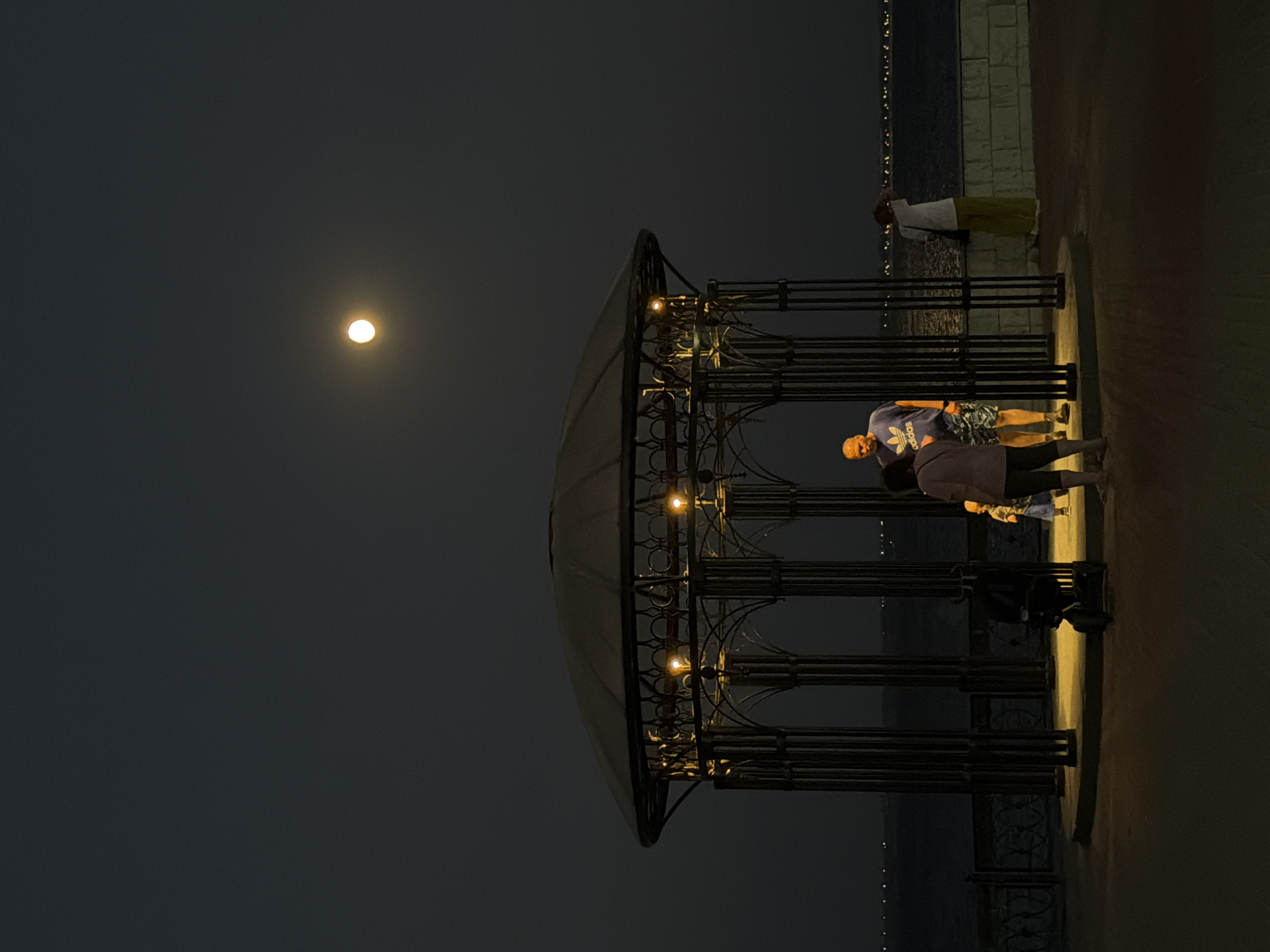 A night scene in Kamyshin, Russia features a gazebo on a promenade near the Volga River. A full moon is visible in the dark sky. Inside the gazebo, a man in a blue t-shirt and patterned shorts stands with a woman in a grey top and dark pants. A child in a blue dress and orange shirt is also present, along with a stroller. A person in a white top and green pants stands near a low wall on the right, looking out at the water. The promenade is lit by the moon and lights within the gazebo, casting yellow light on the ground. The river is calm, with distant lights visible on the opposite bank.