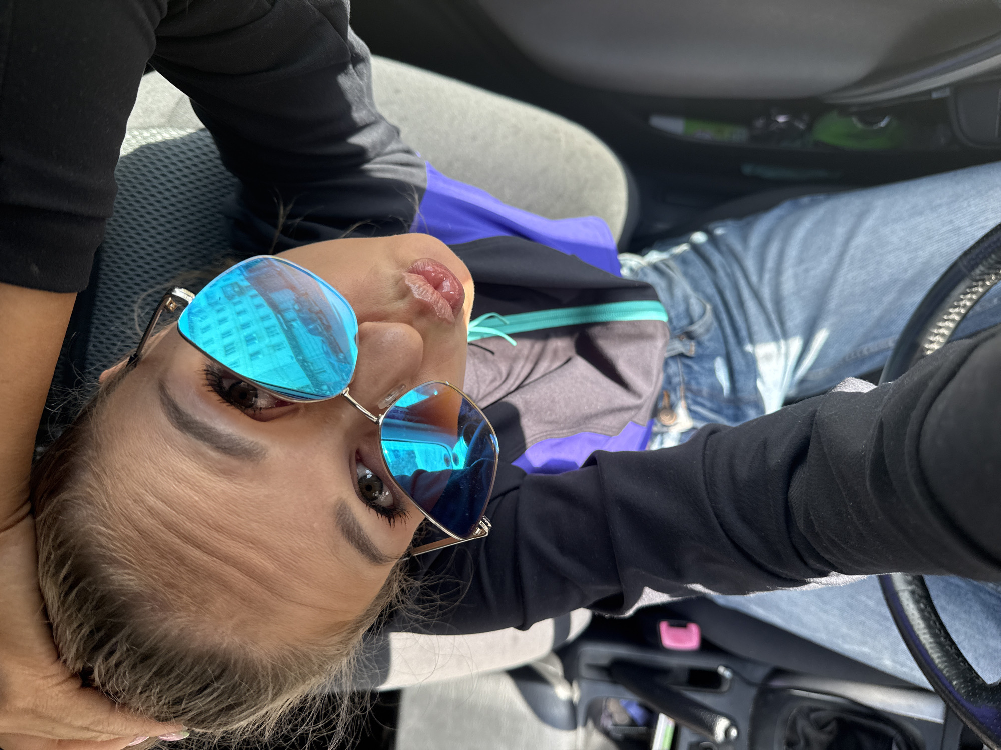 A woman is captured in a high-angle selfie inside a car. She is wearing blue jeans, a black long-sleeved shirt with purple and teal accents, and distinctive blue mirrored sunglasses. Her hair is pulled back, and she is puckering her lips, which are slightly parted. The interior of the car is visible, including a steering wheel, a gear shift, and the car's seating. Sunlight casts shadows, indicating it is daytime. The image's composition suggests the woman is in the driver's seat, holding the phone to take the picture. The overall scene depicts a casual moment, possibly during a drive. There is no identifiable information within the image to place the location in Chaikovsky, Russia.