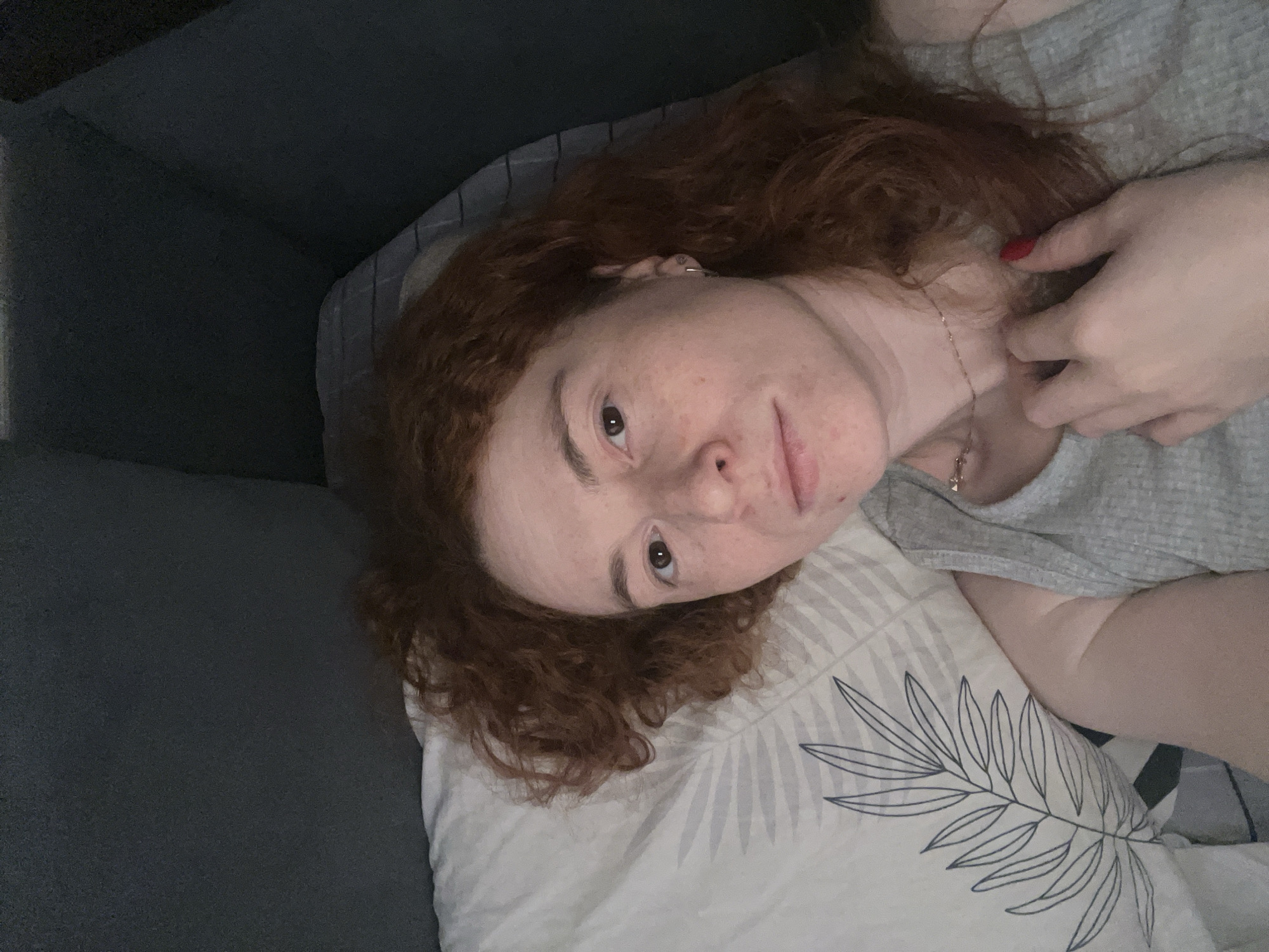 A woman with red curly hair is lying on a pillow with a leafy pattern on a gray couch. She is wearing a gray shirt and a necklace. One hand is visible near her neck, showing red nail polish. The image appears to be taken indoors. The specific location within Chaikovsky, Russia, cannot be determined from the image.
