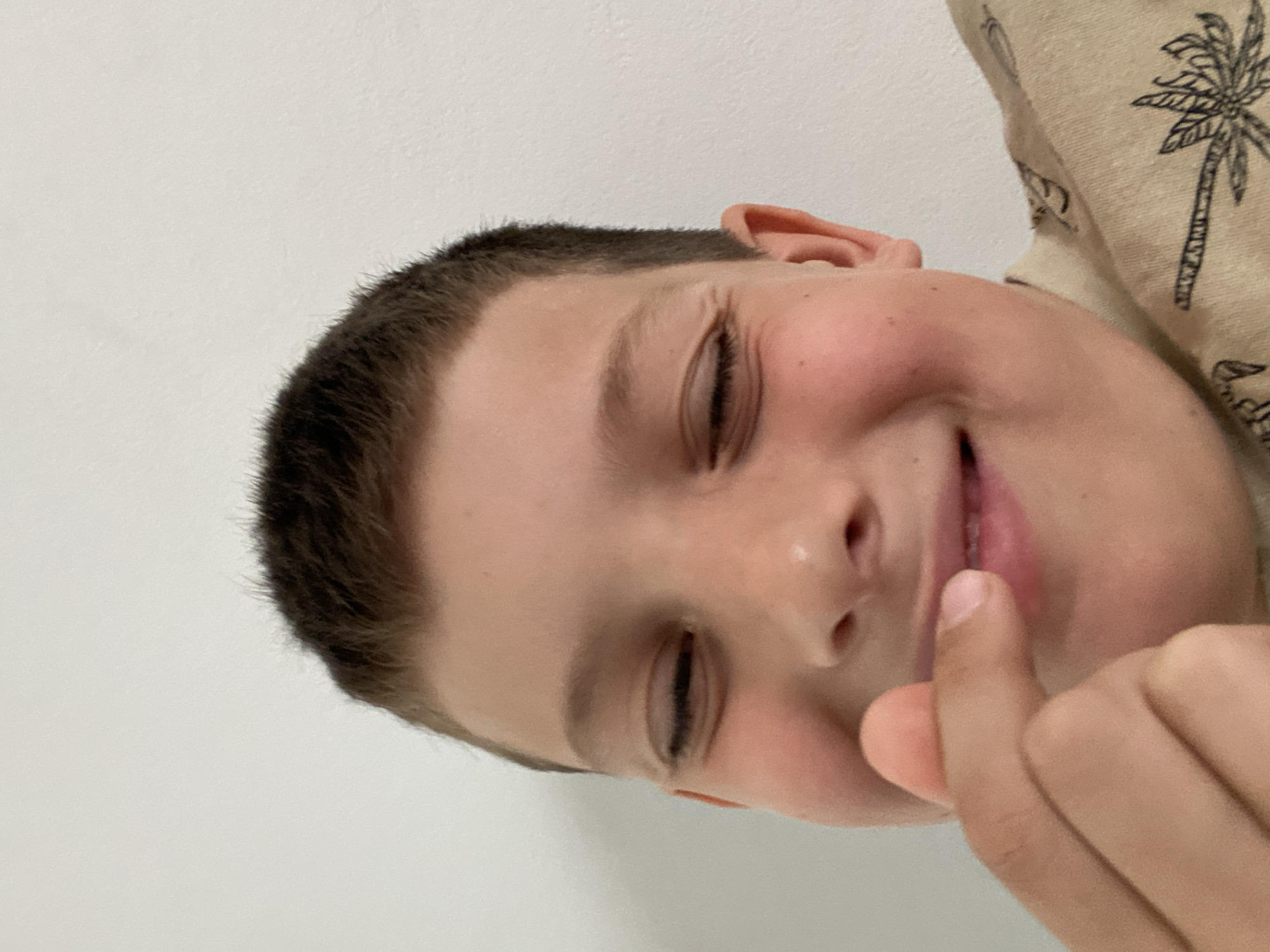 A young child with short brown hair, eyes closed in a possible smile, presses a finger to their lips. The child is wearing a light-colored shirt patterned with black palm trees. The background is a plain white wall. The city of Budva, Montenegro is not discernible.