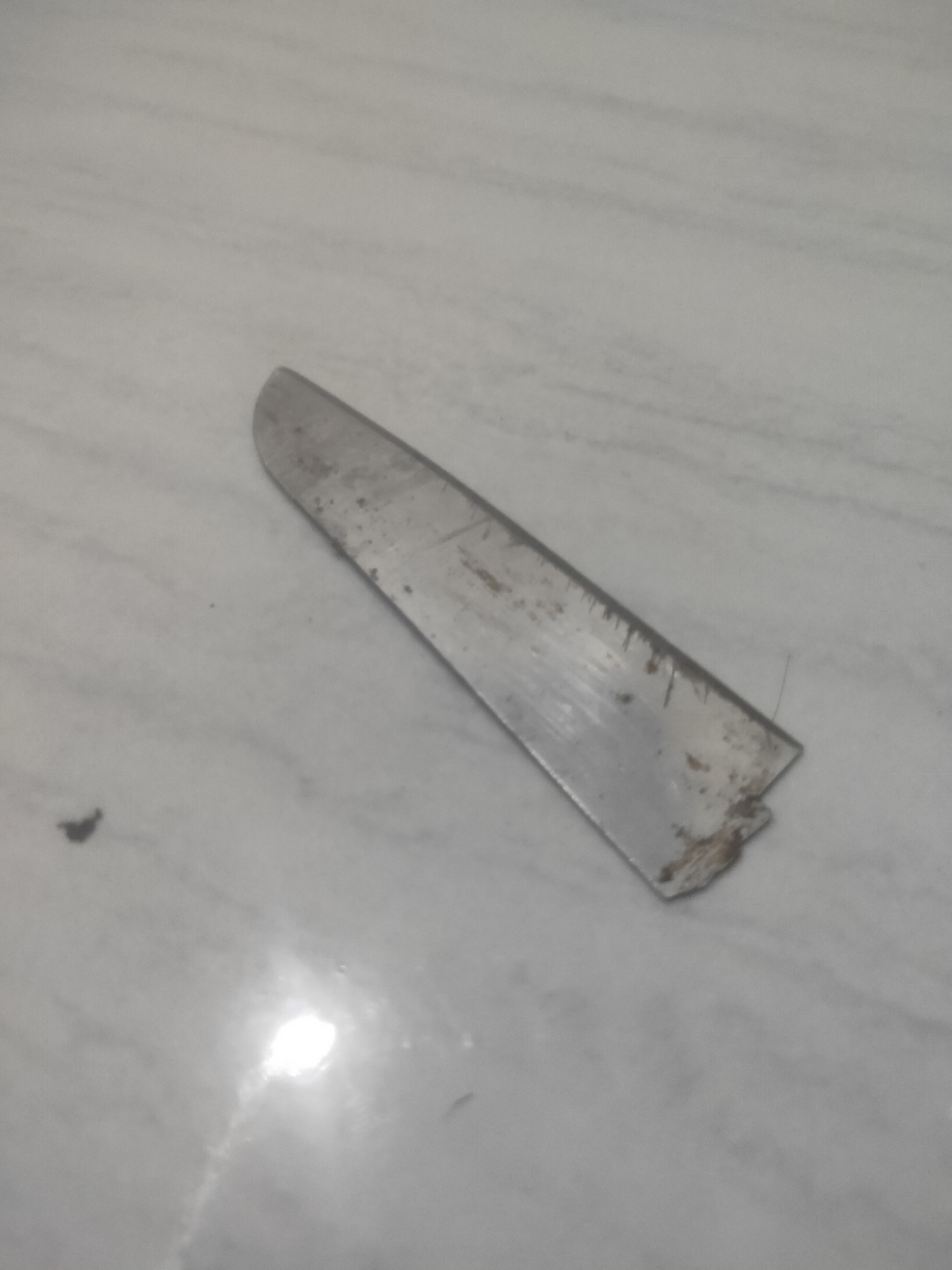 A close-up, overhead shot shows a damaged, dull metal blade lying on a speckled, light-colored surface. The blade is long and flat with a dull edge, exhibiting scratches and discoloration consistent with rust or dirt. The right end of the blade is broken and jagged. The surface appears to be a countertop or tabletop with subtle marbling. The lighting in the scene creates reflections on the surface, obscuring some details. No people or other objects are visible. The location is identified as Antirogo, Indonesia.
