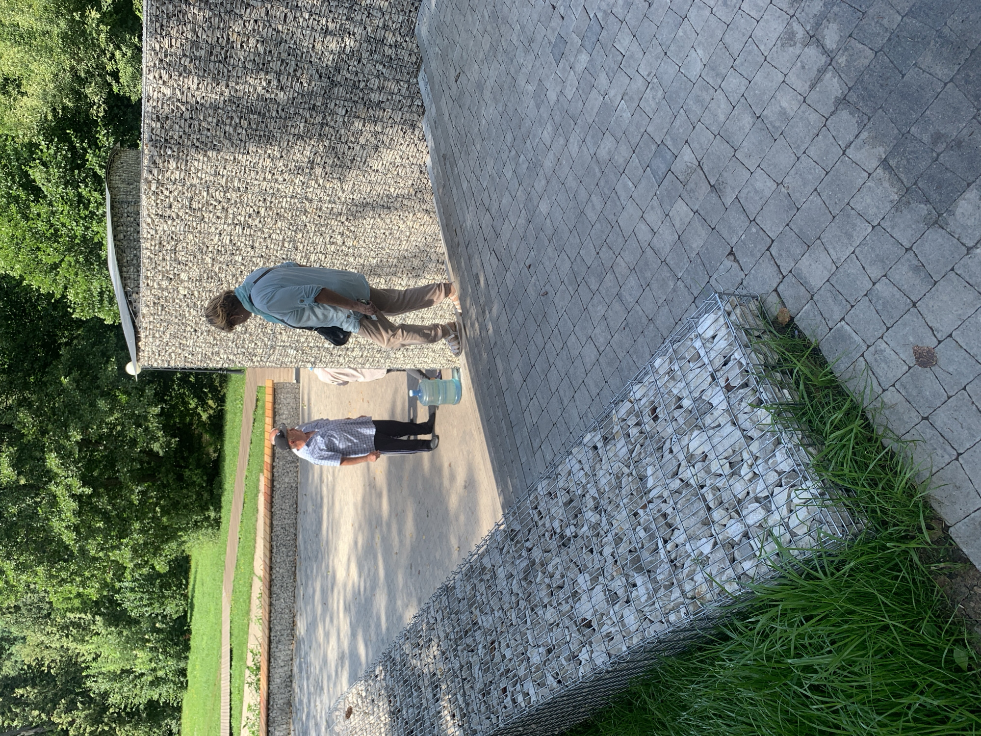 In Arbuzov-Baran, Russia, two men and one woman are present in an outdoor area with paved walkways and a gabion wall. The woman, wearing a light blue shirt and tan pants, is walking down a set of stone steps. To her left, a man in a checkered shirt and dark pants stands near a large water cooler. A second man, partially visible, is standing behind the woman. The gabion wall, filled with light-colored stones, forms a barrier to the right of the scene, and a similar gabion structure is visible on the left, filled with stones and bordering lush green grass. Trees and a grassy area are in the background.