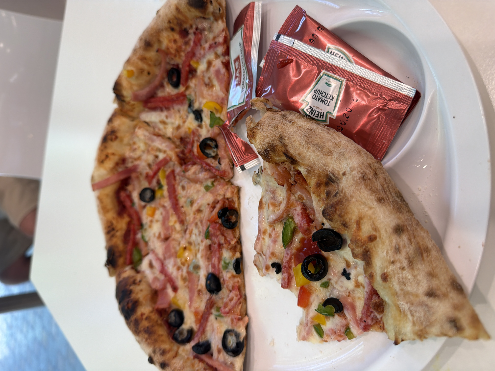 In an indoor setting, likely a restaurant, a white plate holds a partially eaten pizza with a visible crust and toppings of cheese, ham, peppers, and olives. Two packets of ketchup, one Heinz and another with "SAS" branding, are also on the plate. A slice of pizza is placed in the foreground, partially obscuring the ketchup packets. A blurred background shows a person sitting at a table. The scene is lit with natural light. The restaurant is identified as being in Yerevan, Armenia.