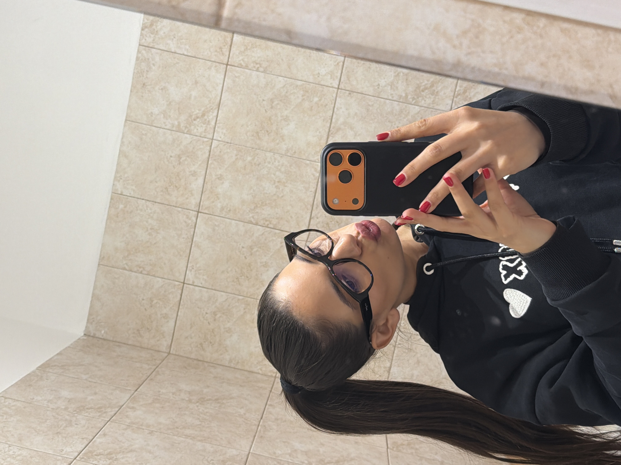 A woman is taking a selfie in what appears to be a bathroom. She is wearing glasses and a black hoodie, and she has her hair pulled back in a ponytail. Her nails are painted red. She is holding a smartphone with a distinctive black and orange case. The background consists of light-colored, square ceramic tiles on the wall and floor. There is a white wall visible. Given the tiled bathroom environment, it is difficult to determine the exact location within Sofia, Bulgaria without further information or visual landmarks.