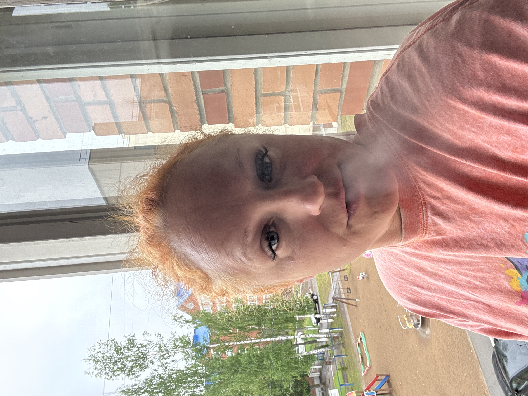 A fair-skinned woman with reddish-blonde hair is looking out a window. The reflection in the window shows a brick building across from her. Outside the window there is a playground and greenery. The playground is located near residential buildings in the city of Chaikovsky, Russia.