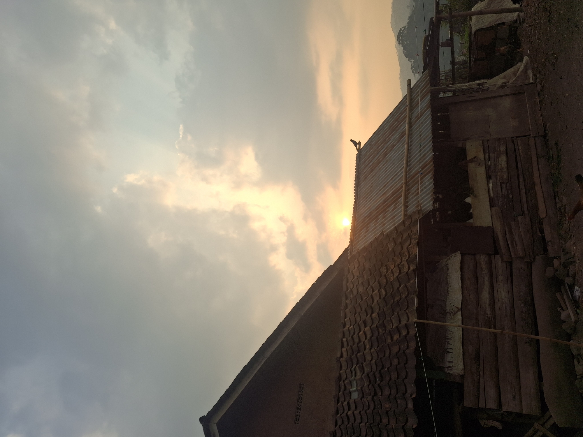 The image depicts a rural scene at sunset in Muncar, Indonesia. A modest, weathered house with a tiled roof dominates the left side of the frame. Adjacent to it is a structure with a corrugated metal roof, possibly a shed or animal enclosure, built from rough-hewn wood. A single bird is perched on the peak of this metal roof. The sky is filled with clouds illuminated by the setting sun, casting a warm, orange glow. In the background, rolling hills are visible under the hazy sky. The foreground shows a dirt ground with scattered debris. The overall impression is one of simplicity and a connection to nature at the end of the day.