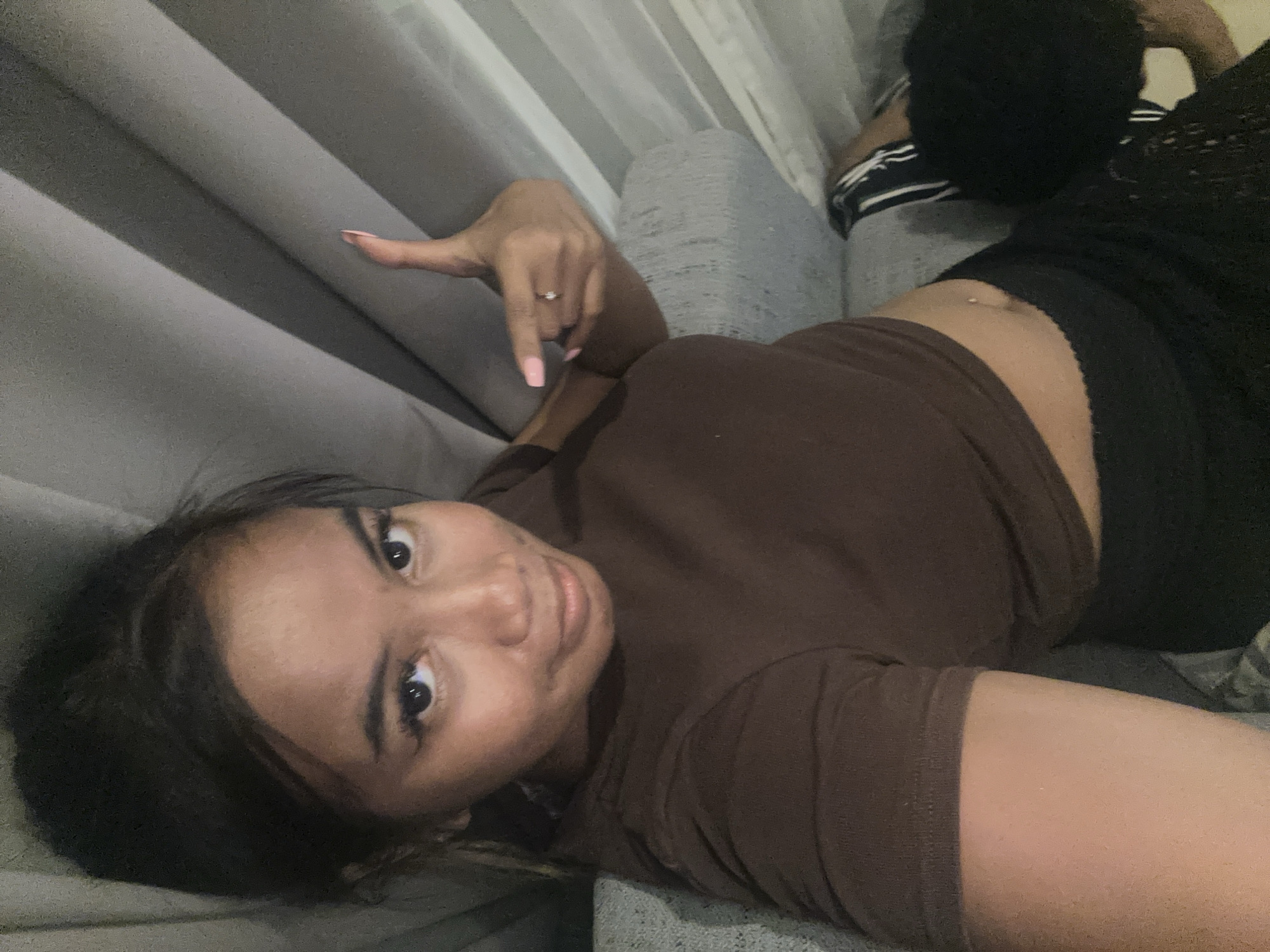 A dark-haired woman is lying on a gray sofa, wearing a brown shirt and black leggings. She has a ring on her left hand. She is looking at the camera. Beside her is a person with black hair. The scene appears to be indoors, possibly a home. The location is Central Jakarta, Indonesia.
