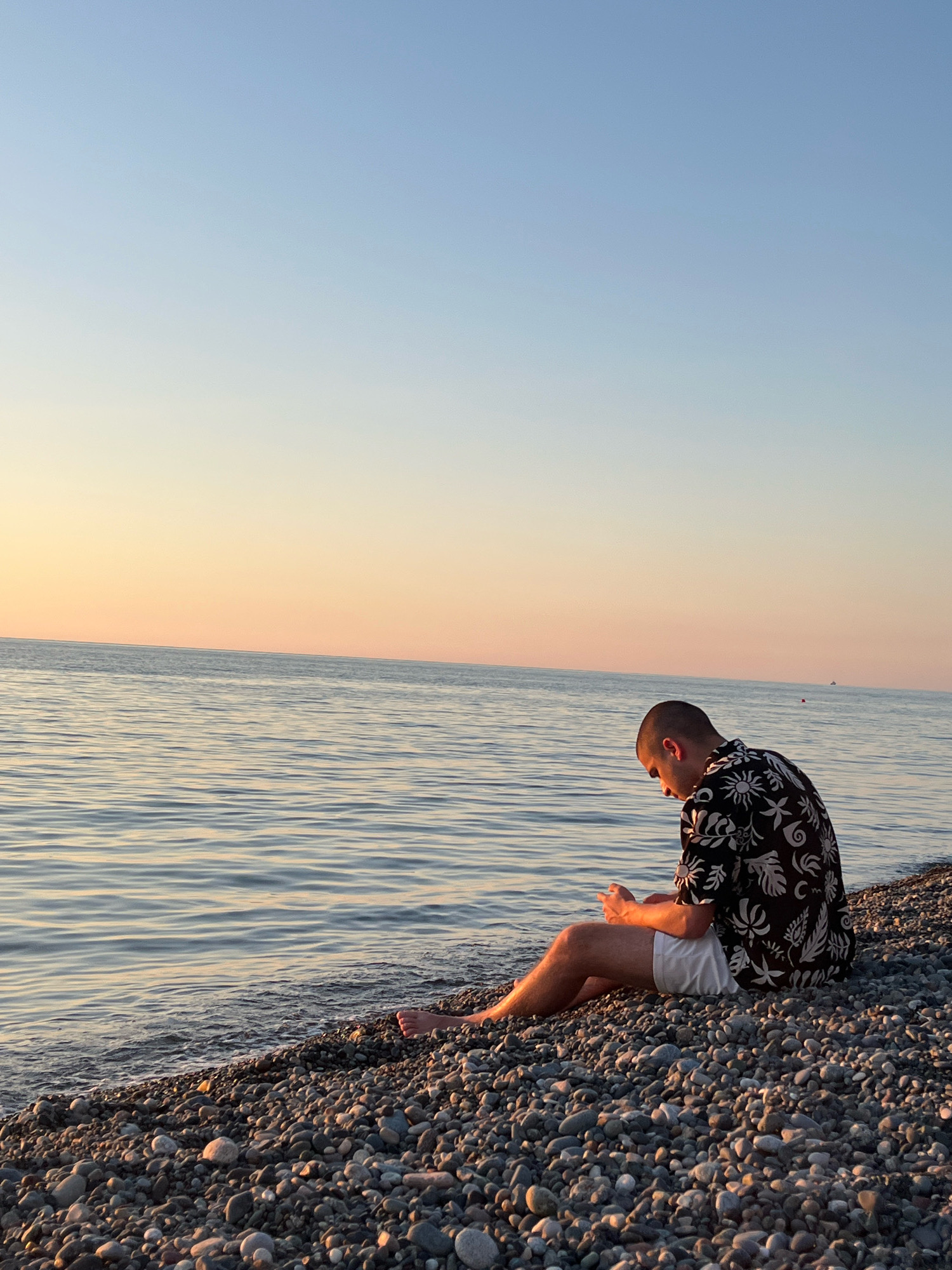 A young man with a shaved head, wearing a black patterned short-sleeved shirt and white shorts, sits on a pebble beach in Kobuleti, Georgia. He is looking down at his hands, which are clasped in his lap. The sea is calm and reflects the soft colors of the sunset sky. The sky transitions from a light blue at the top to a pale orange near the horizon. The pebble beach is covered in smooth, grey stones of various sizes. A few small boats are visible in the distance on the water.