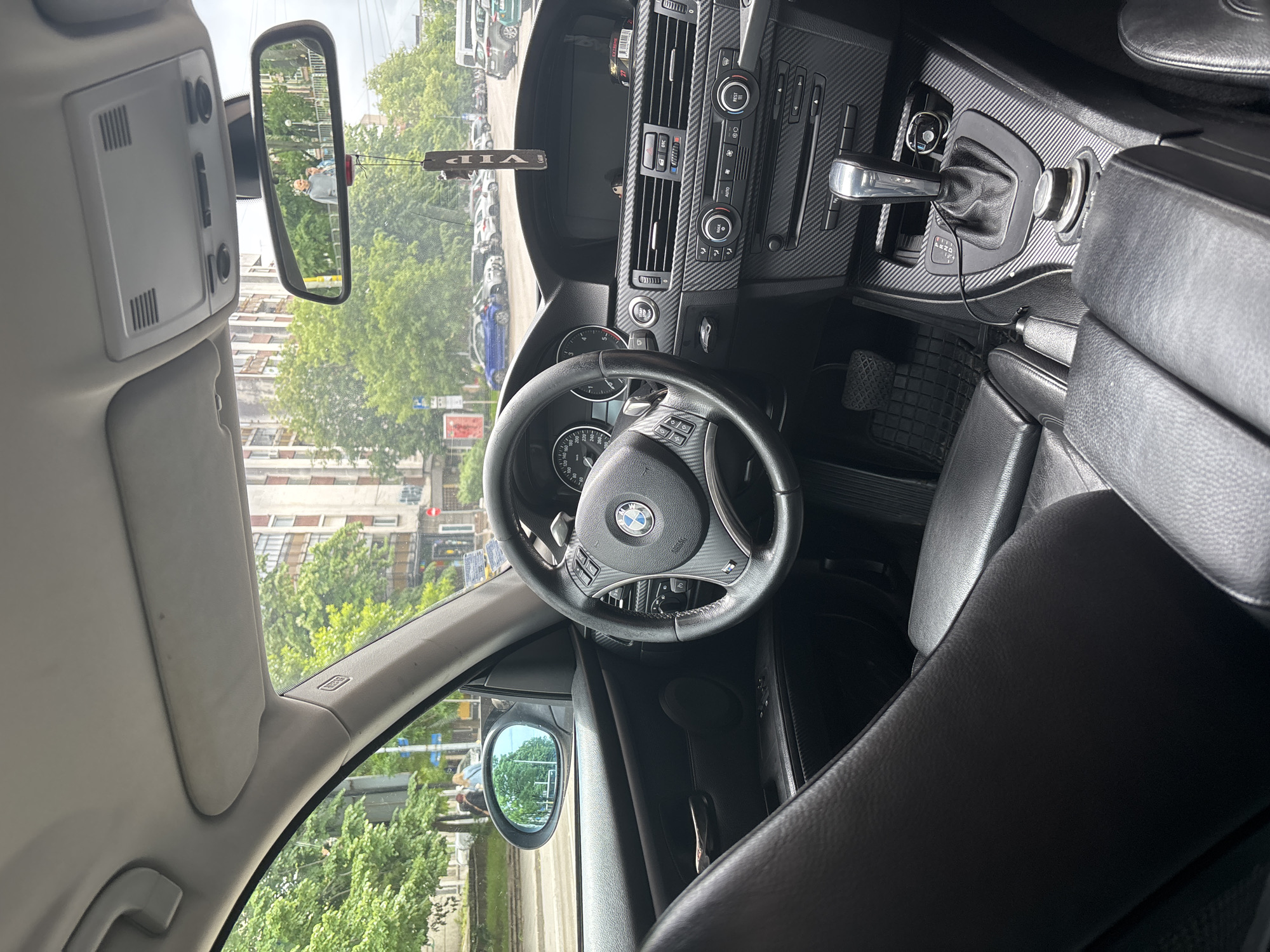 The image depicts the interior of a BMW car, showing the dashboard, steering wheel, seats, and rearview mirror. The dashboard has a carbon fiber-like finish. The steering wheel has the BMW logo in the center. The rearview mirror reflects a street scene with trees, buildings, and pedestrians. The car appears to be parked in an urban area.