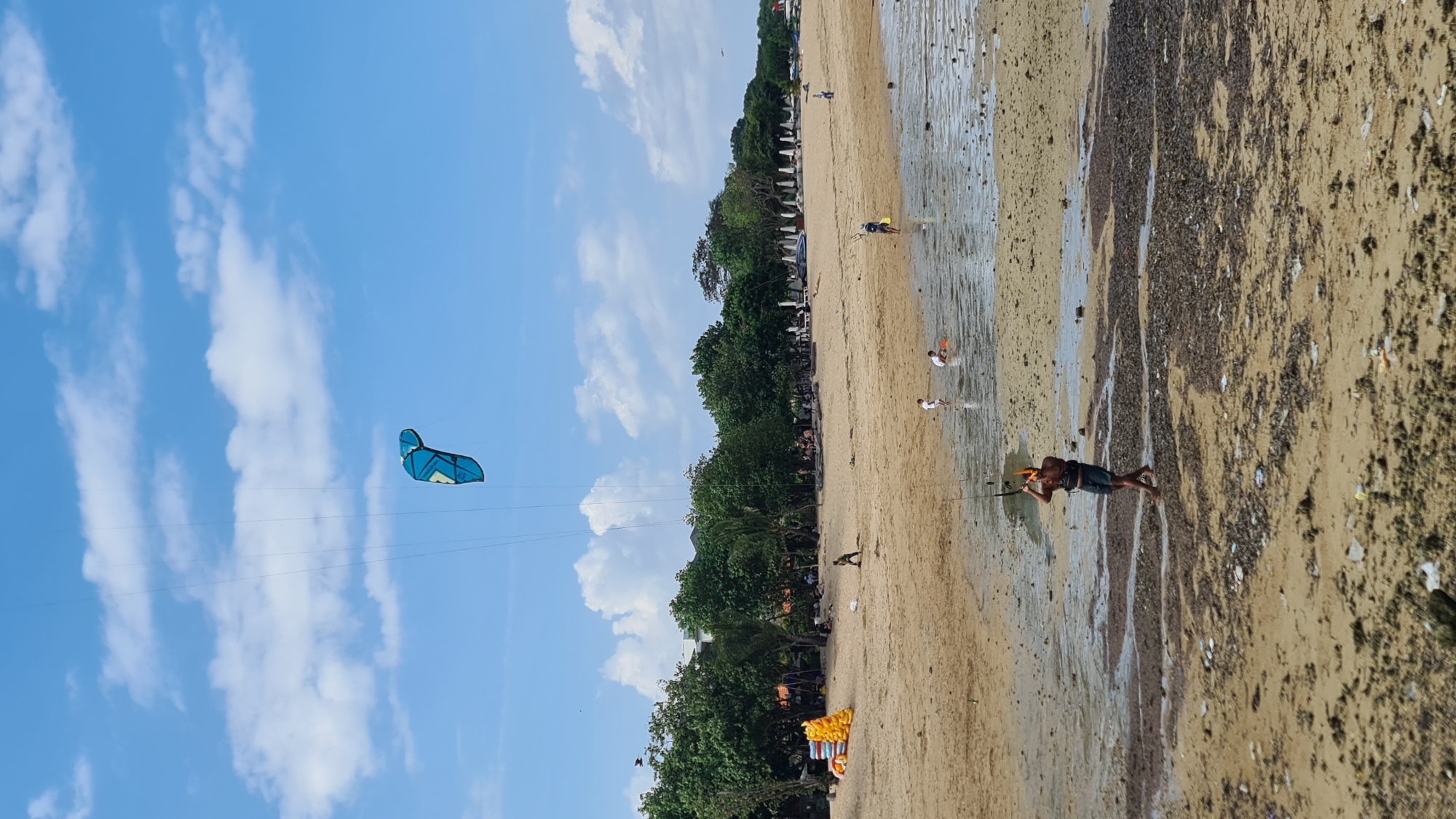 In Denpasar, Indonesia, on a sandy beach, a person is kiteboarding under a bright blue sky with scattered clouds. The kiteboarder is holding onto a control bar, maneuvering a blue and yellow kite high in the air. The beach is populated with a few other individuals, some wading in the shallow water and others relaxing further back near a line of trees and umbrellas. To the left, several yellow inflatable objects are stacked on the sand, suggesting a rental or activity area. The tide appears to be low, revealing wet sand and shallow pools on the beach.