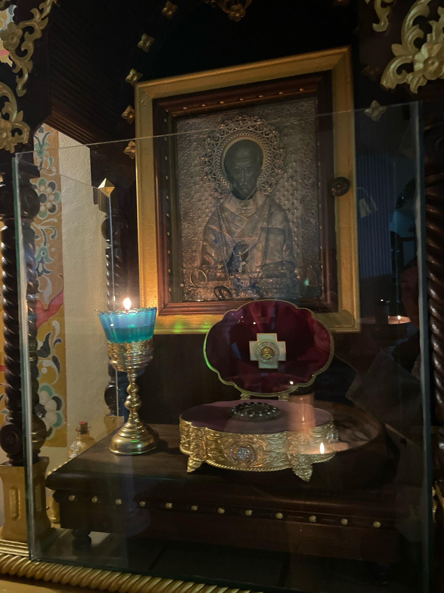 The image displays a religious altar within a glass enclosure, likely in Pattaya City, Thailand. The central focus is an icon of a male figure, framed and adorned with intricate metallic detailing. To the left of the icon, a lit blue glass candle holder with a golden base holds a flickering flame, casting a warm glow. Below the icon, a velvet-covered pedestal displays a golden cross with a central medallion, set against a rich, dark background. The altar is situated on a dark wooden cabinet, which is itself placed on a decorative golden base. The background shows decorative elements of an interior, with ornate woodwork and patterned walls. The scene is captured from a slightly angled perspective, emphasizing the reverence and solemnity of the religious display.
