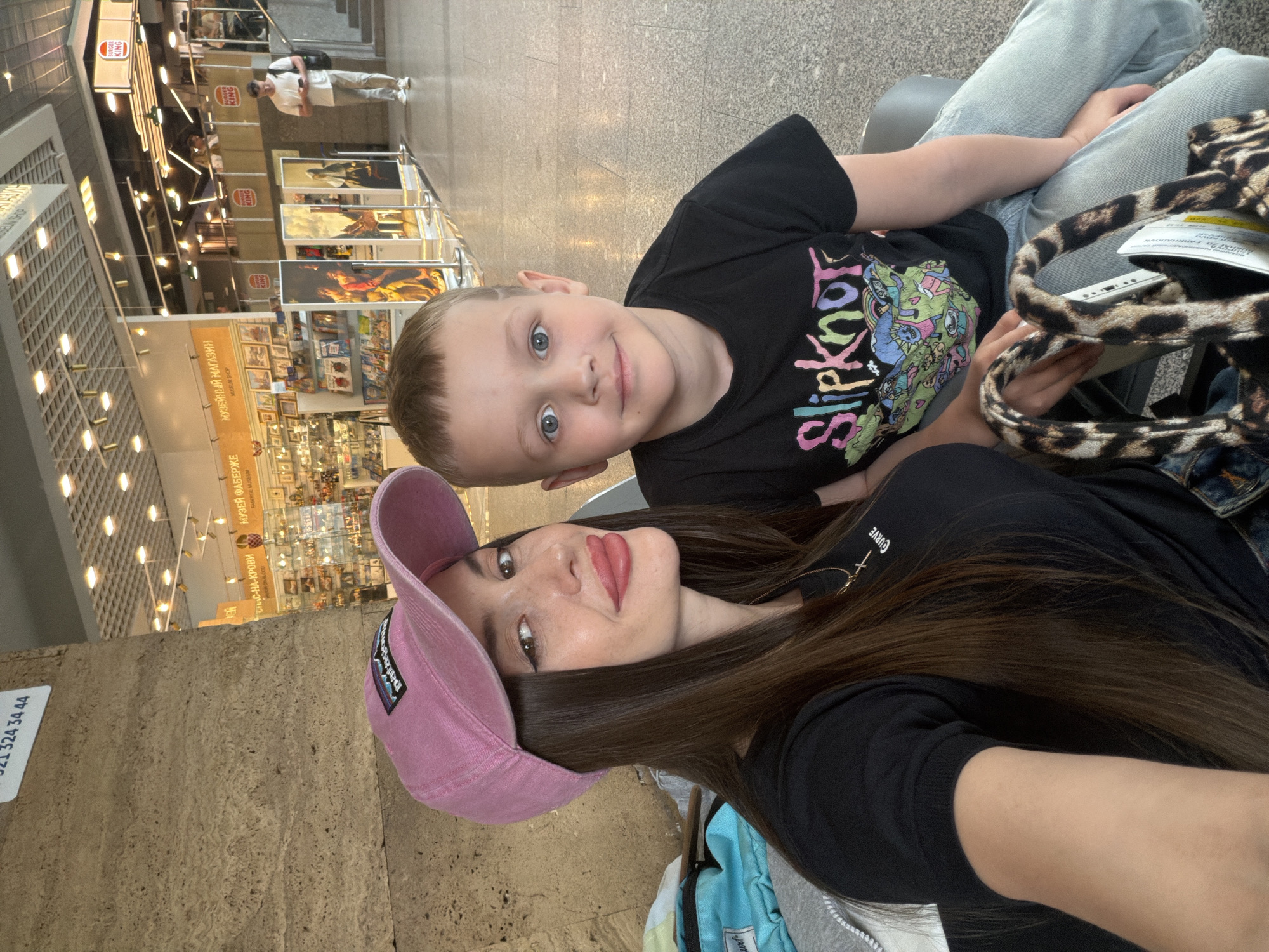 The image captures a selfie featuring a woman and a young boy, both smiling, in an indoor public setting, likely a commercial or cultural complex. The photo is rotated 90 degrees counter-clockwise.
The woman, positioned closer to the camera, wears a black t-shirt with "CURVE" printed vertically and a pink baseball cap featuring a "Patagonia" logo. She has long, dark brown hair and is wearing red lipstick and eyeliner. A delicate cross pendant hangs from her neck. The boy, next to her, has short blonde hair and blue eyes. He is wearing a black t-shirt with a colorful "Slipknot" graphic. Both are looking directly at the camera with pleasant expressions, suggesting a relaxed moment.
Visible objects include a leopard print bag held by the woman and a light blue backpack. The background indicates a busy environment with bright artificial lighting. Multiple "BURGER KING" signs are visible, alongside cultural references. Signs in Cyrillic read "МУЗЕЙ ФАБЕРЖЕ" (FABERGE MUSEUM) and "МУЗЕЙНЫЙ МАГАЗИН" (MUSEUM SHOP). Another sign partially visible says "СПАС-НА-КРОВИ" (Church of the Savior on Spilled Blood), strongly suggesting the location is in Saint Petersburg, Russia. Other individuals are seen walking in the distance, and various displayed items or artworks are visible in the shop areas. A stone-textured pillar or wall on the left side shows a partially obscured sign with numbers "324 34 44". The scene suggests the pair is seated, possibly waiting, in a well-lit public area.