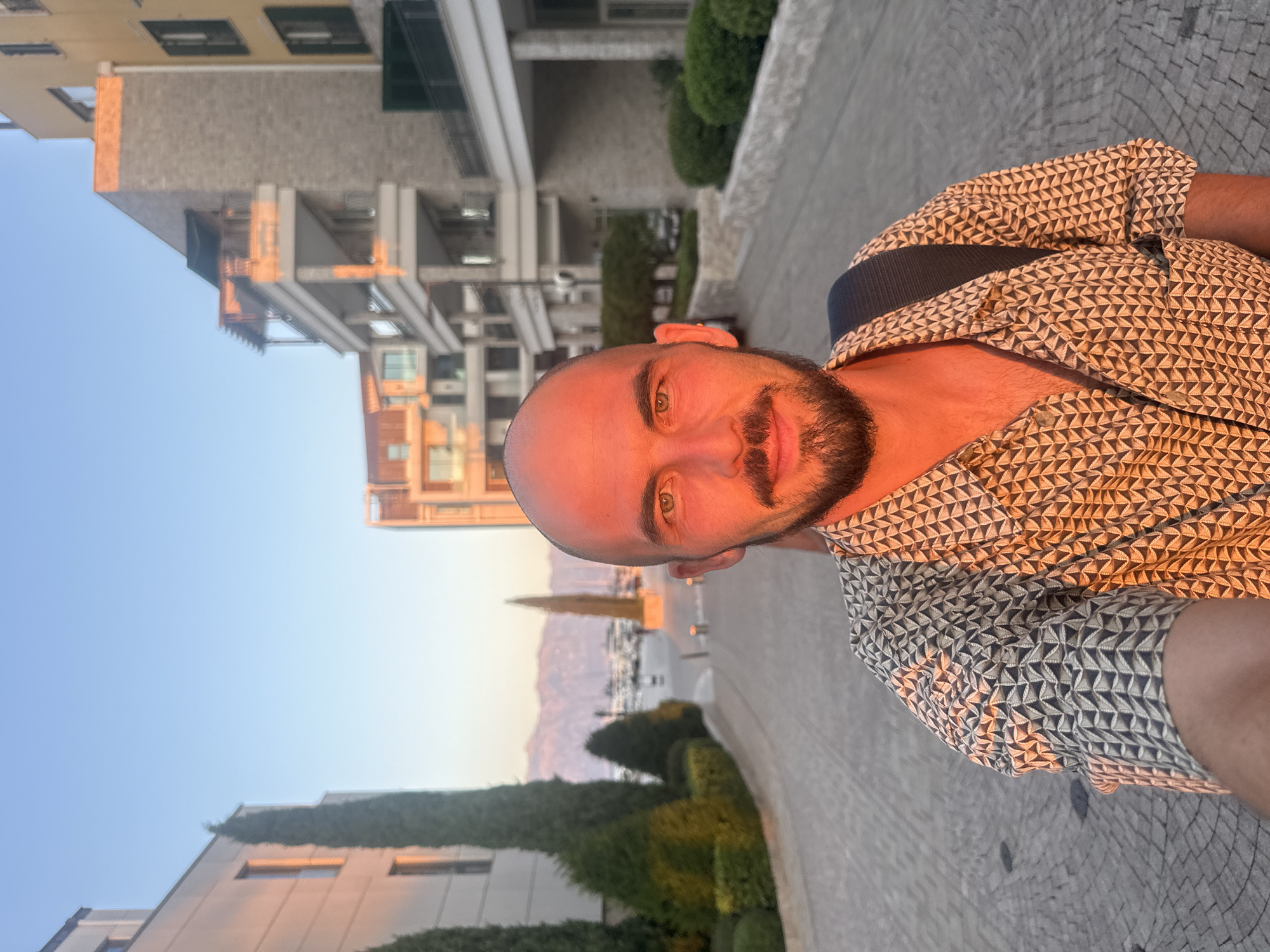 A man with a shaved head and a beard takes a selfie on a street in Kumbor, Montenegro. He is wearing a patterned short-sleeved shirt and a backpack. The street is paved with cobblestones, and lined with manicured bushes and trees. In the background, there are modern apartment buildings with balconies, and beyond that, a body of water with boats and mountains in the distance. The sun is setting, casting a warm orange glow on the scene.