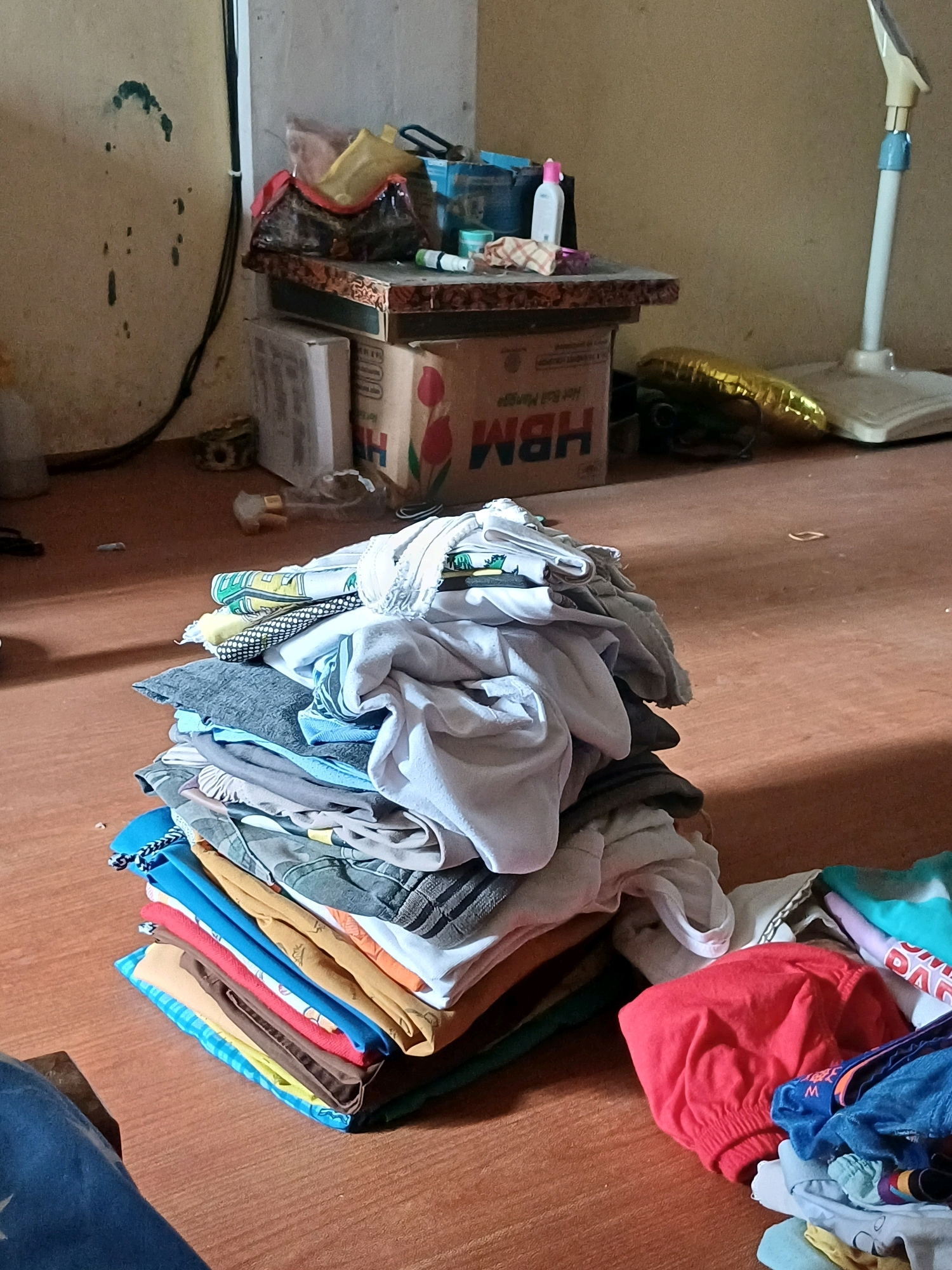 A pile of folded clothes sits on a wooden floor in what appears to be a room. The clothes are various colors and types, including t-shirts, pants, and possibly towels. Behind the clothes, there is a small table with some items on it, including a box, bags, and a bottle. To the right of the table, there is a standing fan. The walls of the room are a light color and appear to be somewhat worn. The overall impression is that of a domestic setting, possibly a bedroom or living room. There is no clear indication of the specific location within Dumai City, Indonesia, beyond it being an indoor scene.