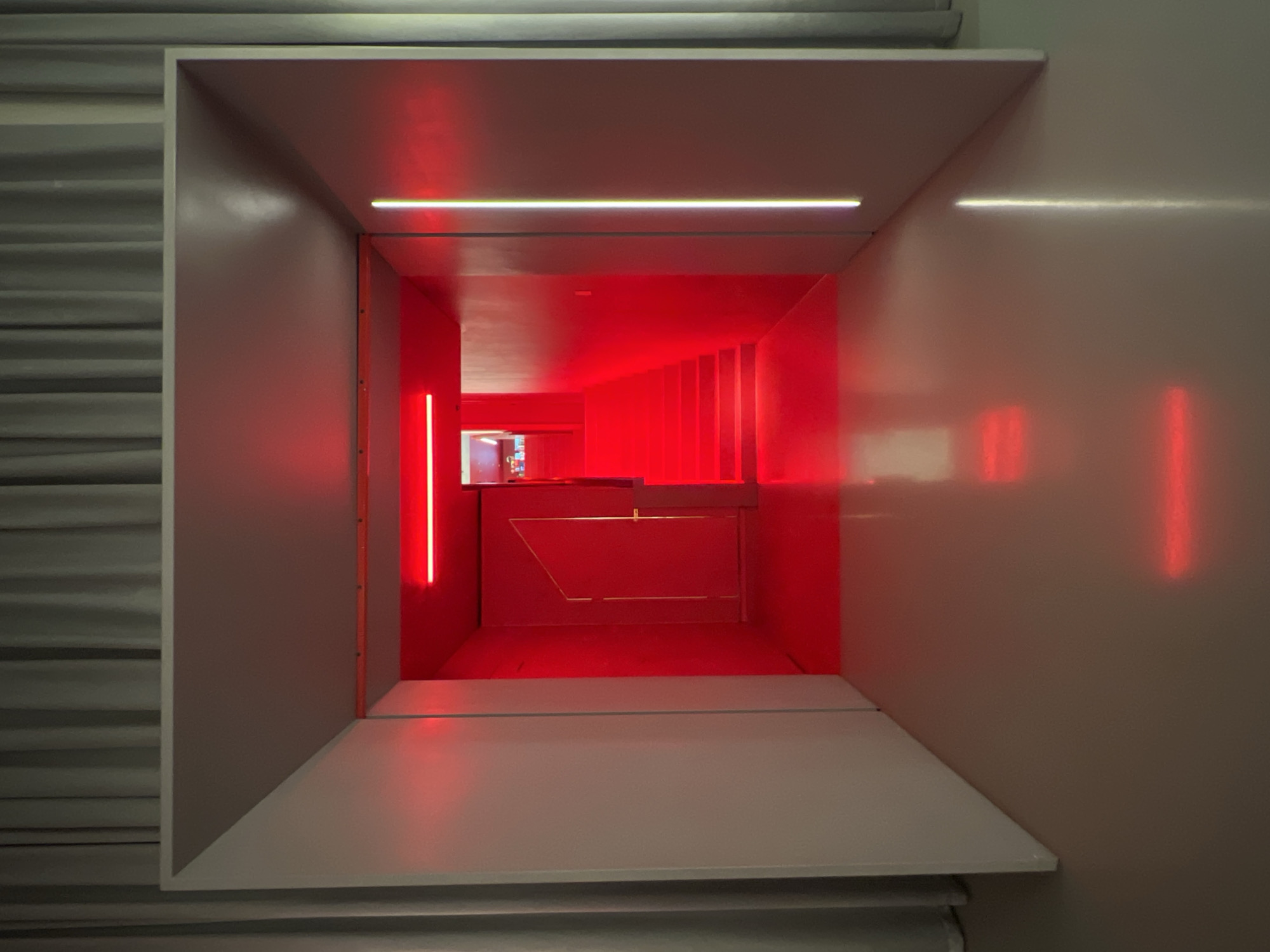 The image presents a rectangular, tunnel-like opening illuminated by red light. The external structure is composed of gray or white panels, with horizontal lines visible on the side, perhaps indicating corrugated metal. The red light emanates from within the tunnel, highlighting vertical elements and a desk or counter-like surface. The opening is framed by a flat, horizontal surface in the foreground, which could be a shelf or platform. The red lighting scheme suggests a modern or artistic installation. The location is not discernible from the image.
