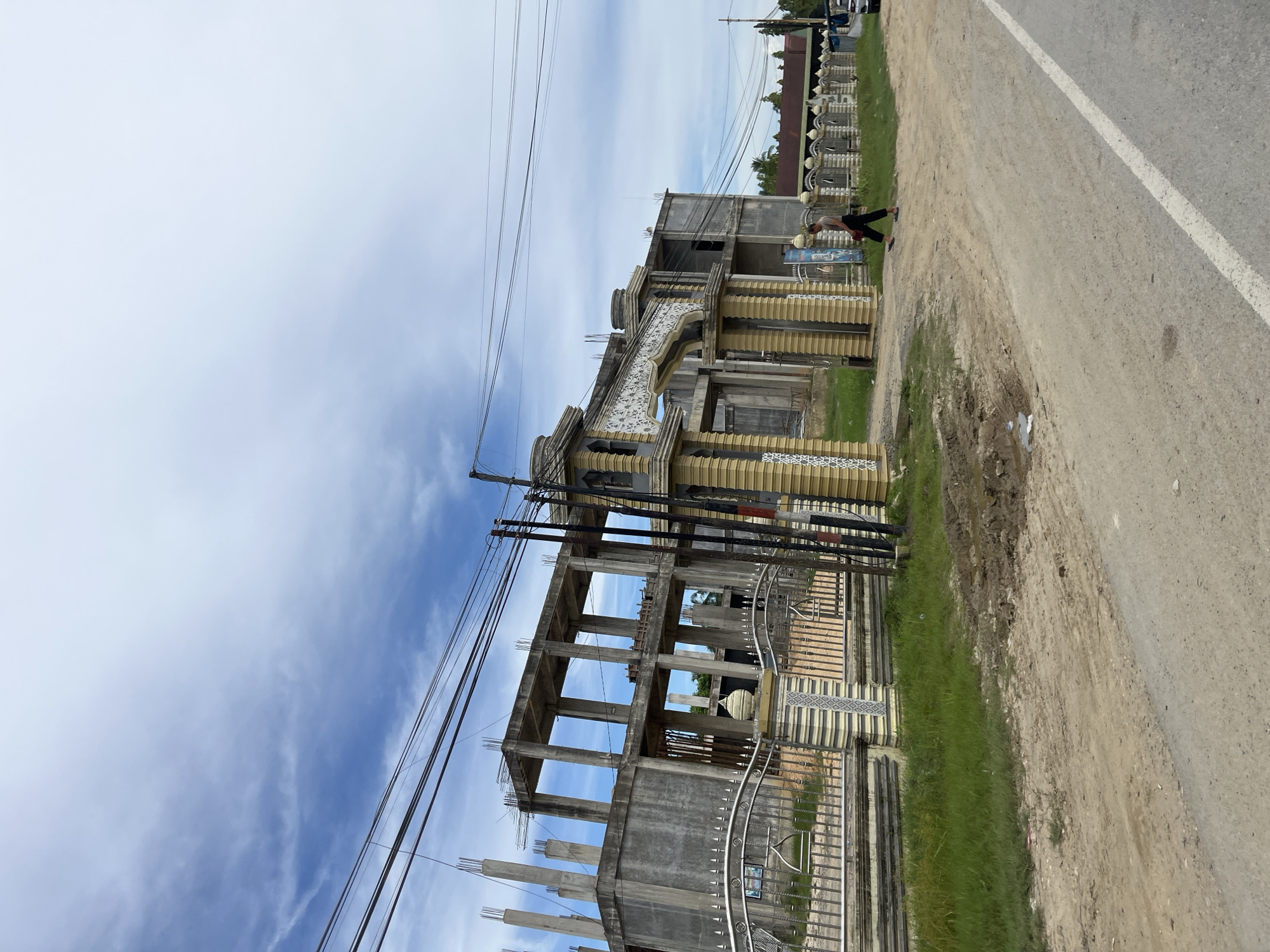 A full shot captures an unfinished concrete building with exposed rebar on the upper floors, located in Simpangempat, Indonesia. The building is fenced with a decorative metal and stone wall, featuring an ornate archway. A lone individual walks across a dirt path in front of the building. The surroundings are characterized by a dirt road, grassy areas, and utility poles with numerous wires. The sky is partly cloudy.