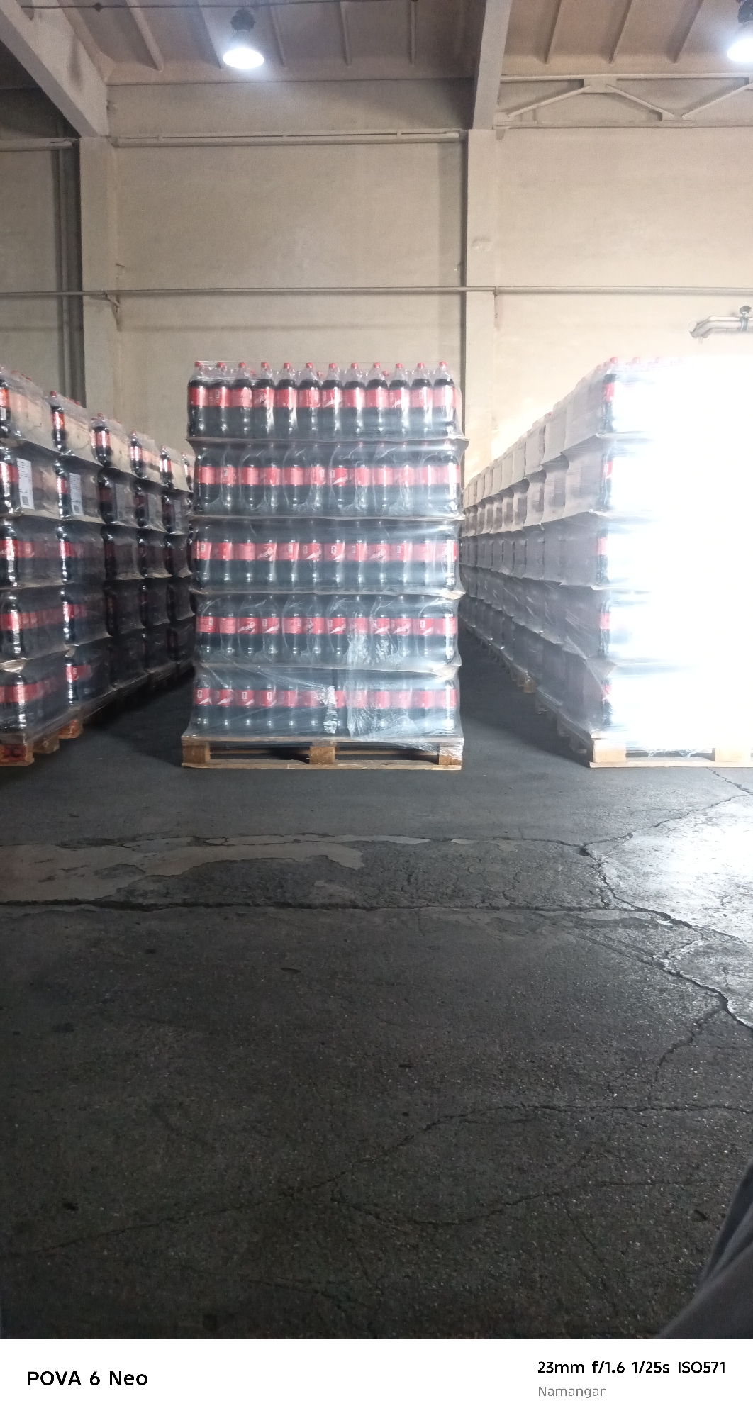 The image shows a warehouse interior in Namangan City, Uzbekistan, with several stacks of bottled beverages, likely Coca-Cola due to the distinctive red and black labeling visible on the bottles. The bottles are shrink-wrapped onto wooden pallets, creating tall, rectangular stacks. Two large stacks are prominently featured in the center of the image, with more stacks extending into the background on the right. The warehouse appears to be well-lit by overhead industrial lighting. The floor is a dark, cracked asphalt or concrete surface. The overall scene depicts a storage or distribution area for a large quantity of bottled drinks.