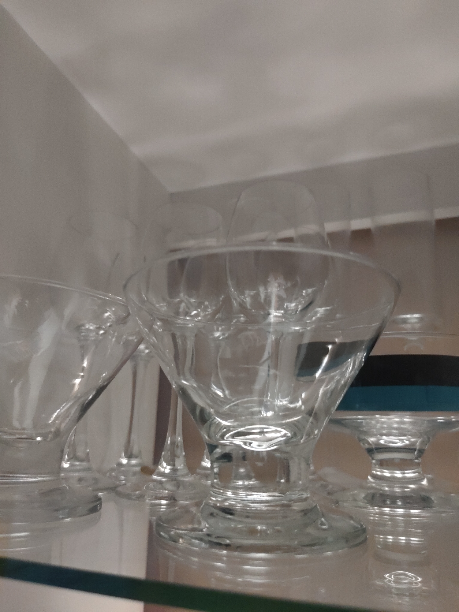 The image depicts a collection of clear glass stemware arranged on a reflective surface within an enclosed space, likely a cabinet or display unit, in Davao City, Philippines. The foreground features a prominent martini-style glass with a wide, conical bowl and a short stem, surrounded by several other clear stemmed glasses of varying designs, including taller wine glasses and additional cocktail or dessert glasses. One glass on the right exhibits a distinct dark blue band around its lower section. The glassware rests on a highly reflective shelf, mirroring their bases. A green horizontal line is visible at the bottom of the frame, marking the edge of the shelf or a foreground element. The background consists of plain light gray or off-white walls, with a visible corner in the upper left indicating the interior of a confined space. The scene is illuminated by soft, diffused light, producing gentle highlights and reflections on the glass. No individuals are present, and no actions or interactions are depicted.