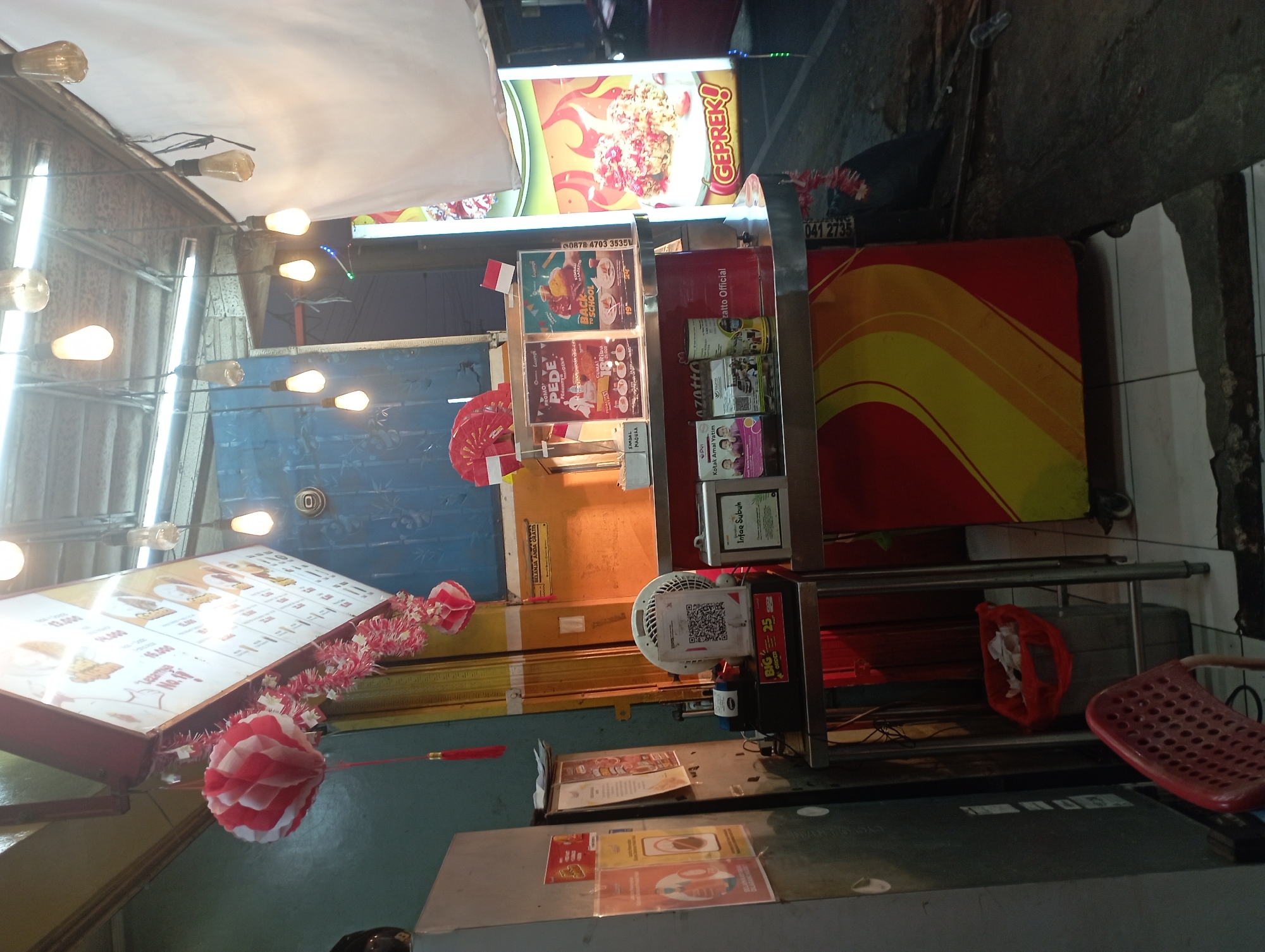 This image depicts an outdoor food stall in Purwakarta, Indonesia, likely at night given the artificial lighting. The stall features a red and yellow awning and is illuminated by several hanging light bulbs and a brightly lit sign. The sign displays an image of food with the word "GEPREK!" in large, bold letters, suggesting a spicy chicken dish. A menu board with food items and prices is visible above the counter.
Various objects are present at the stall, including a small fan, a QR code for payment, and several promotional flyers. A red chair is placed in front of the stall, and a red trash can with some discarded items is visible to the left. The background shows a blue wall and a yellow metal structure, possibly part of a building or another stall. The overall scene is indicative of a small business operating in a bustling urban or semi-urban environment.