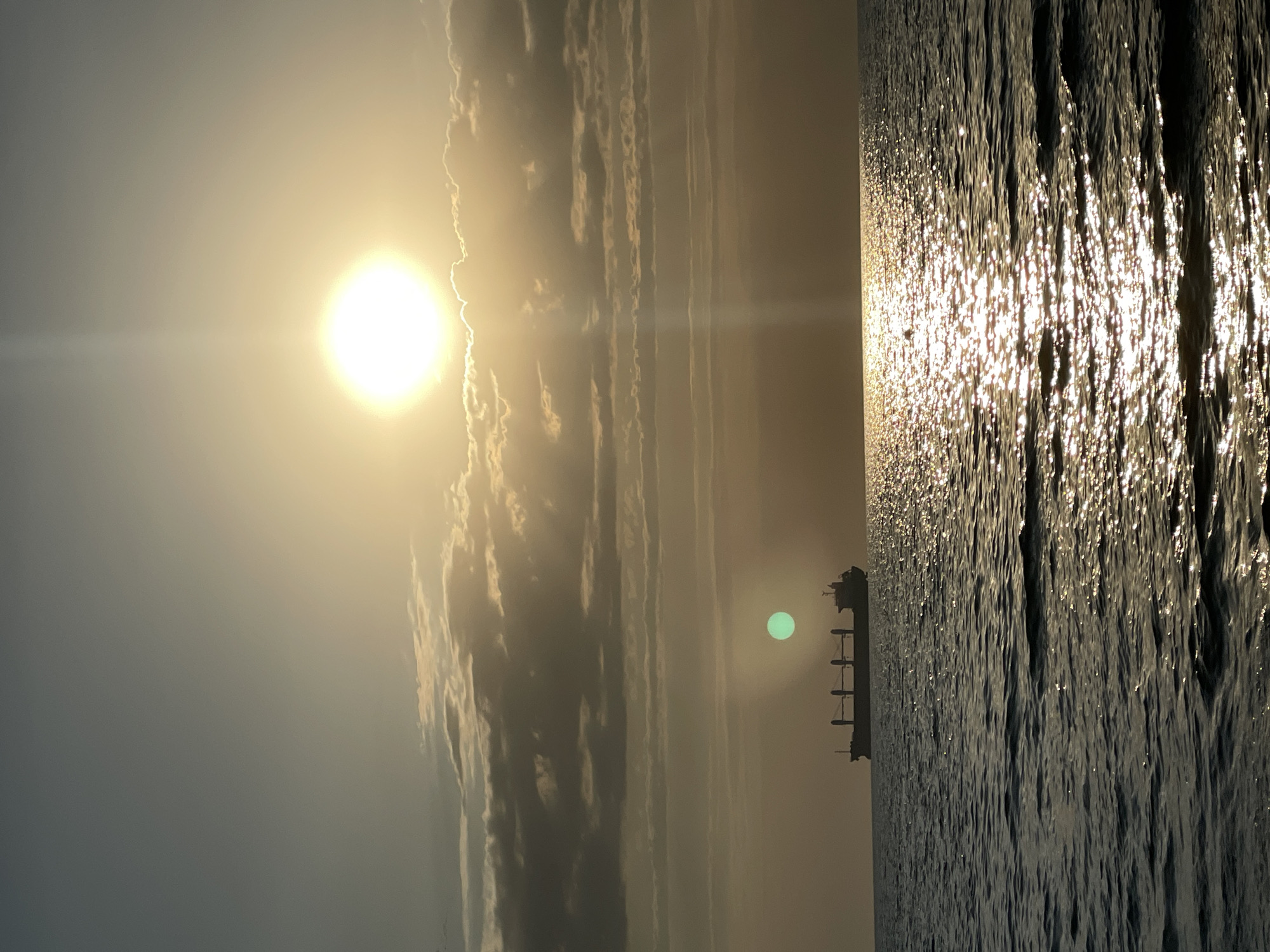 An aerial shot captures a large cargo ship sailing on the sea, silhouetted against a bright sun. The sun is partially obscured by clouds, creating a halo effect and a beam of light extending downwards. The sea is choppy with sunlight reflecting off the waves, creating a sparkling effect. The sky transitions from a light blue at the top to a hazy yellow near the horizon. A faint green orb is visible in the sky to the left of the ship. The scene is indicative of a sunset or sunrise over the Black Sea, likely near Anapa, Russia, given the context provided.