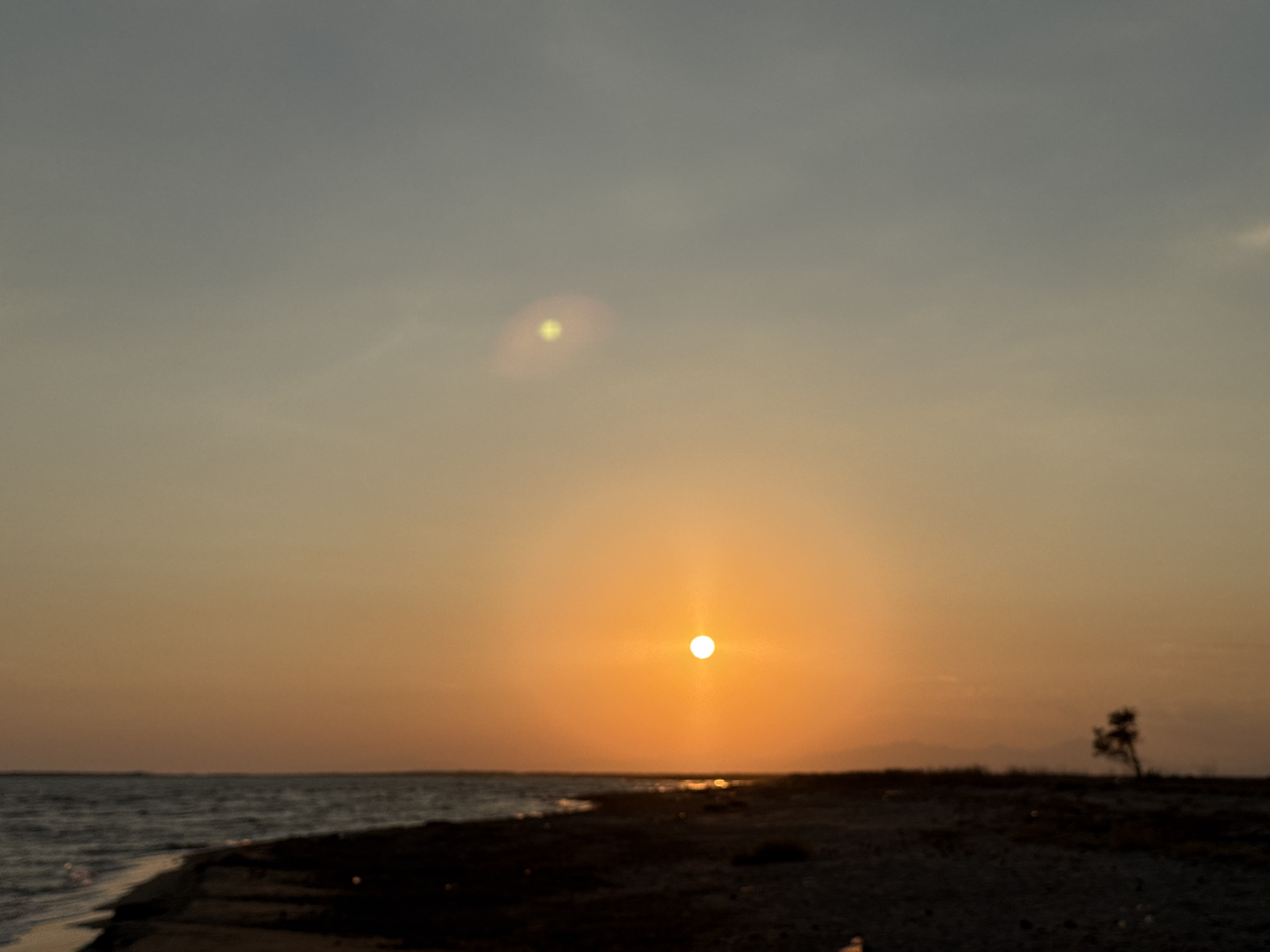 The image depicts a blurry sunset over a body of water, likely a sea or large lake, with a dark, sandy shoreline in the foreground. The sky transitions from a pale blue at the top to a warm orange and yellow near the horizon where the sun, a bright white disc, is positioned. A faint lens flare appears in the upper portion of the sky. The shoreline is mostly dark and appears to be undeveloped, with some sparse, low-lying vegetation. A lone, silhouetted tree stands in the distance on the right side of the frame. In the far distance, faint mountain ranges are visible against the horizon. The water is dark and appears calm with gentle waves lapping at the shore. The overall impression is one of tranquility and natural beauty. There are no visible people or man-made structures in the immediate vicinity. This scene is consistent with a coastal location such as the shores near Imeros, Greece, which is known for its natural landscapes and sunset views.