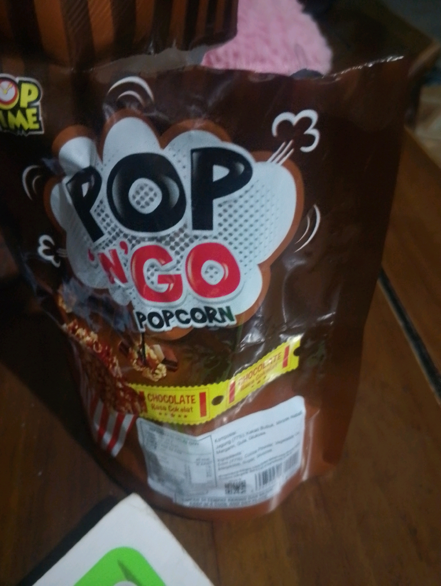 The image shows a close-up of a brown bag of "POP N' GO" popcorn, specifically the chocolate flavor. The bag is lying on a wooden surface, possibly a table. The lighting is dim, and the focus is on the product packaging. The bag features a cartoon-like design with bold lettering and graphic elements. A yellow label on the bag indicates "CHOCOLATE Rasa Cokelat." In the background, there is a hint of a pink fuzzy object and some vertical striped pattern on the top left of the bag. The image does not contain any identifiable people or specific city landmarks.