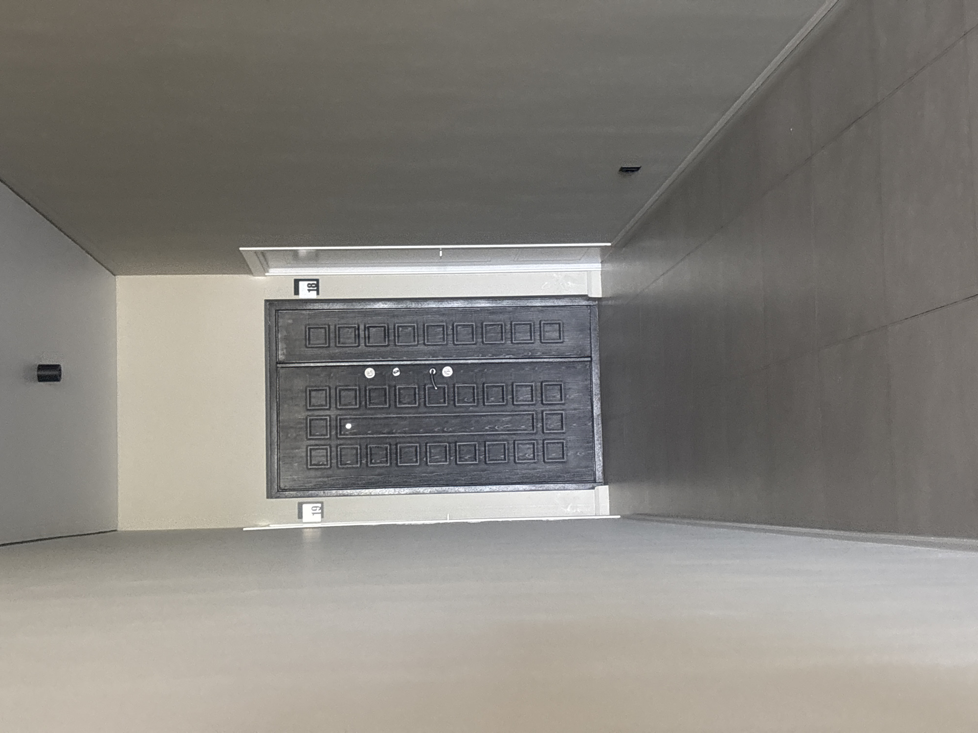 The image shows a narrow, dimly lit corridor with a plain, neutral color scheme. The walls are painted in a light, matte finish, and the floor is tiled with large, grey square tiles. The corridor extends into the distance, with a single light source visible at the far end, creating a long shadow.
In the center of the image, a dark, heavy-looking double door is visible. The doors have a textured surface with a grid of square indentations. On the wall to the left of the door, a small, white illuminated sign displays the number "19". To the right of the door, a similar sign displays the number "18". Adjacent to the door marked "18" is a narrow white doorframe, suggesting another entrance or room.
The overall impression is one of a functional, unadorned interior space, possibly within a residential or commercial building. There are no people visible in the image. The environment appears clean and orderly, but devoid of any decorative elements or personal touches. The lighting is subdued, creating a somewhat austere atmosphere.