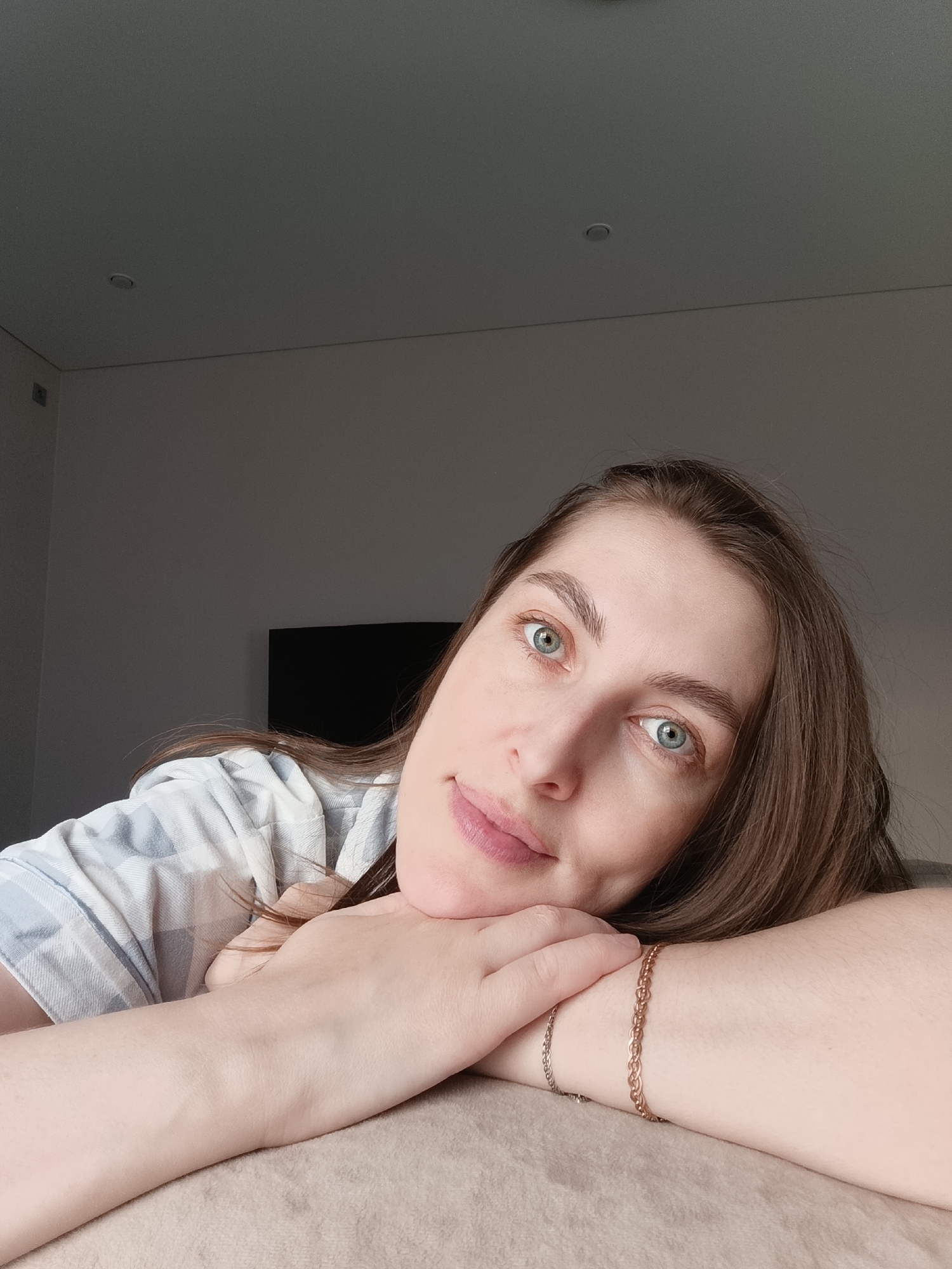 A woman with long brown hair and blue eyes is lying down with her head resting on her hands. She wears a light blue and white striped shirt. A gold bracelet is visible on her right wrist. The background shows a wall with a television screen. The location is in a room in a building in the city of Chaikovsky, Russia.
