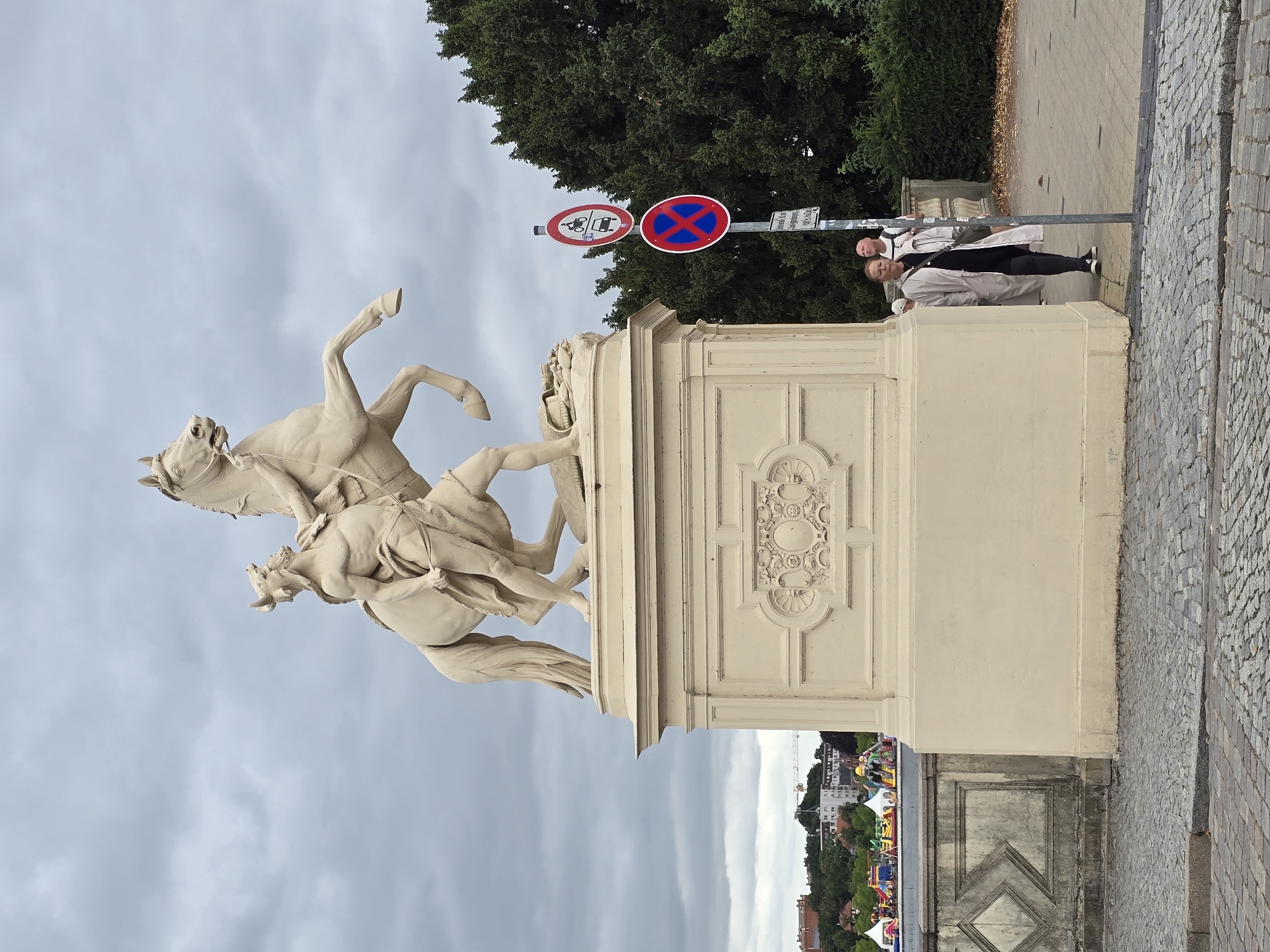 An outdoor, eye-level, full shot captures a stone equestrian statue dominating the center. The statue depicts a muscular man with a beard and horns, holding a sword and reins, mounted on a rearing horse. The statue rests on a large, ornate, cream-colored pedestal with decorative carvings. To the right of the statue, a paved walkway is visible, lined with lush green trees. Two individuals are walking on the walkway; one woman in a light-colored trench coat and dark pants, and a man with a shaved head behind her. A metal pole with traffic signs is positioned between the statue and the walkway. The background reveals a city skyline with buildings and a body of water. A cloudy, gray sky completes the scene. The cobblestone ground in the foreground suggests an urban setting. Based on the equestrian statue and architecture, this image appears to be located in Schwerin, Germany, possibly on a bridge or promenade.