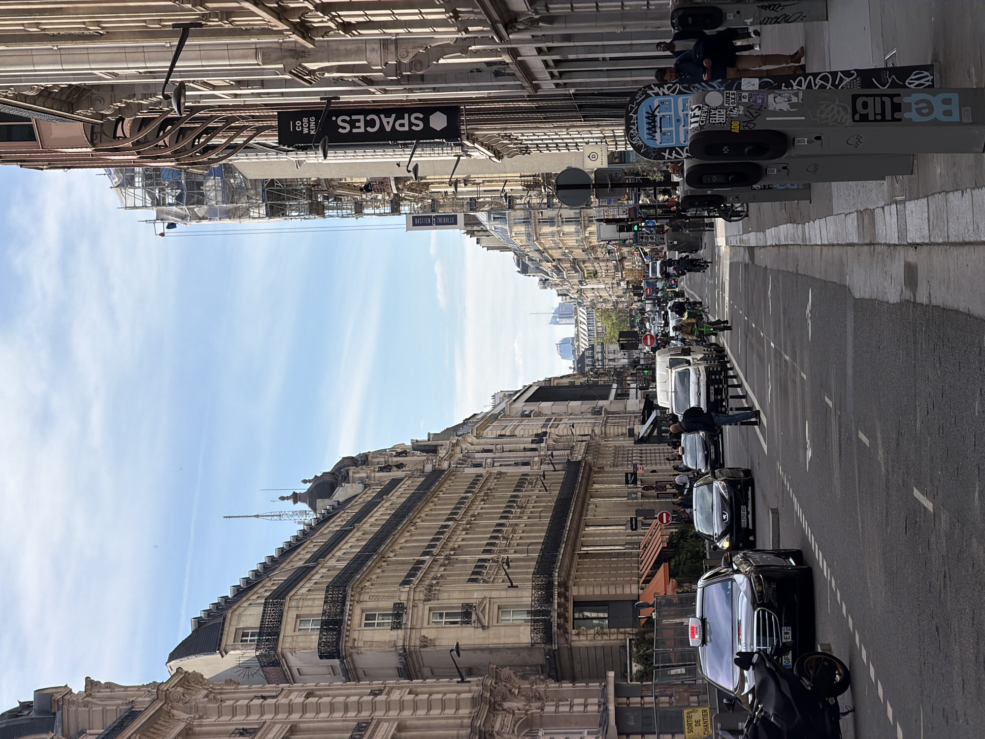 The image depicts a street in Paris, France, viewed from street level, with buildings lining both sides of the road. The architectural style of the buildings is classic Parisian, featuring detailed facades, ornate balconies, and mansard roofs. Scaffolding covers some structures, indicating ongoing construction or renovation work.
The street is busy with traffic, including cars, taxis, motorcycles, and vans. People are walking and standing on the sidewalks, near bicycle-sharing stations, which display the "Belib" logo.
Signs such as "SPACES. CO WORKING" and "BASTIEN ET TREBULLE" are visible on the buildings. There are also traffic signals and road markings indicating lanes. The sky is blue with some clouds visible in the distance, suggesting a bright day.