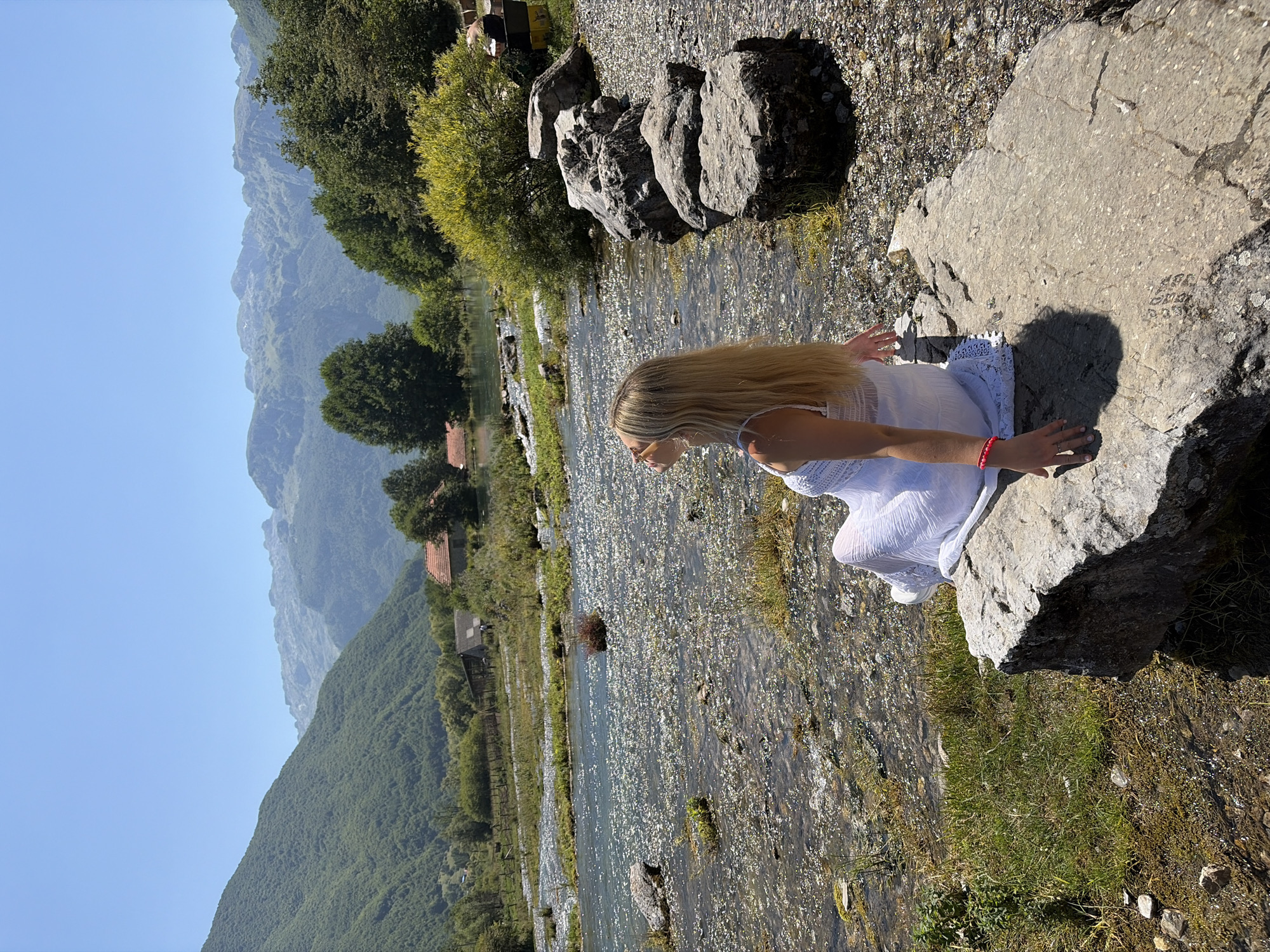 A blonde woman wearing a white dress is sitting on a large rock next to a flowing river. The woman is facing away from the camera, looking towards the mountains in the background. The river is shallow and clear, with stones visible on the riverbed. In the background, there are green, tree-covered mountains and a clear blue sky. The scene appears to be in a rural area. The location is likely Gusinje, Montenegro, known for its mountainous landscape and rivers.