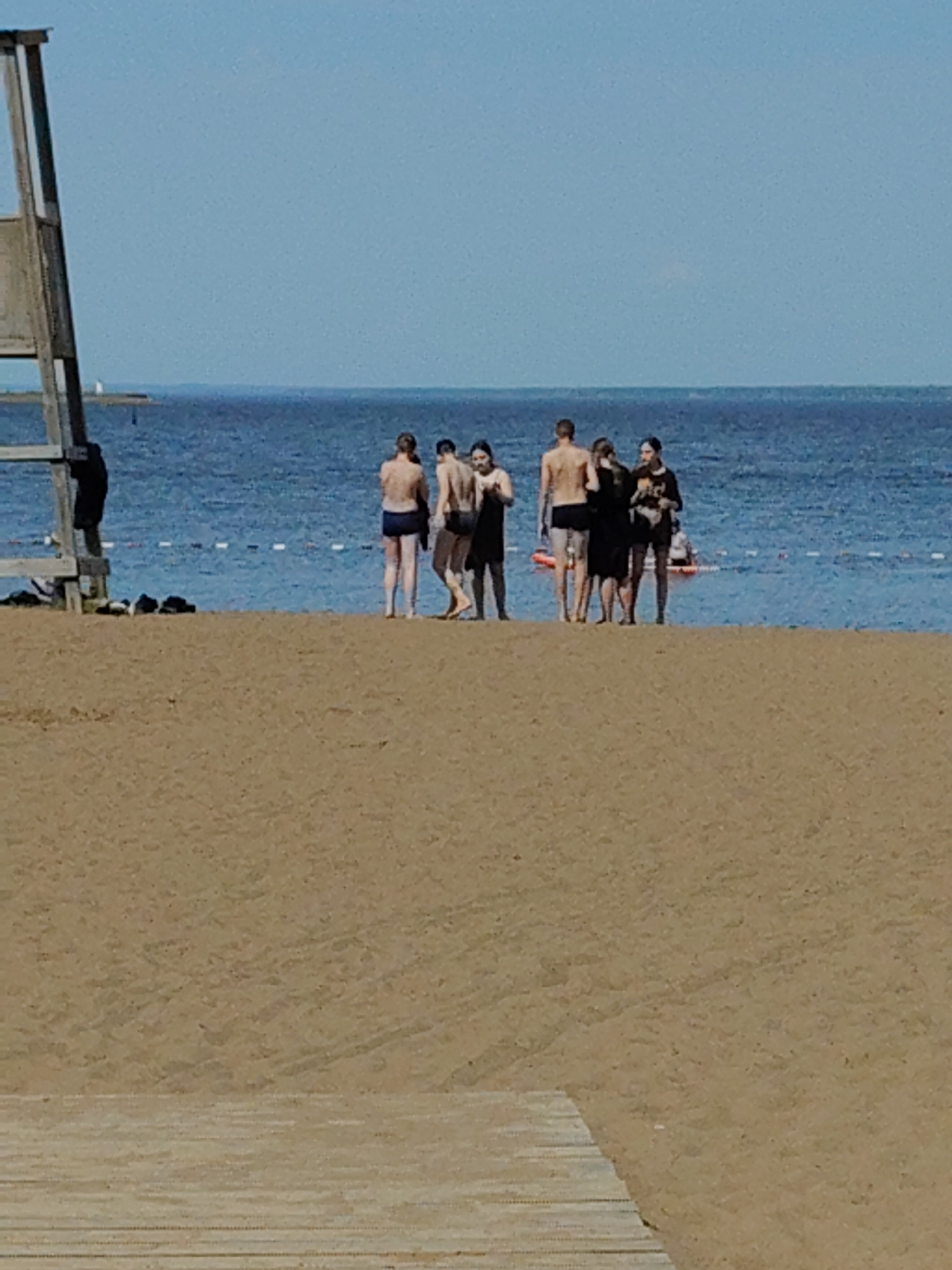 The image captures a group of seven people standing on a sandy beach near the water's edge in the city of Unknown, Unknown. The water appears calm with a line of white floats visible. The sky is clear and blue. To the left of the group, there is a lifeguard stand. The foreground shows a sandy beach with wooden planks visible at the very bottom of the frame. The people seem to be of varying ages, dressed in swimwear and casual clothing. They are standing together, some in conversation.