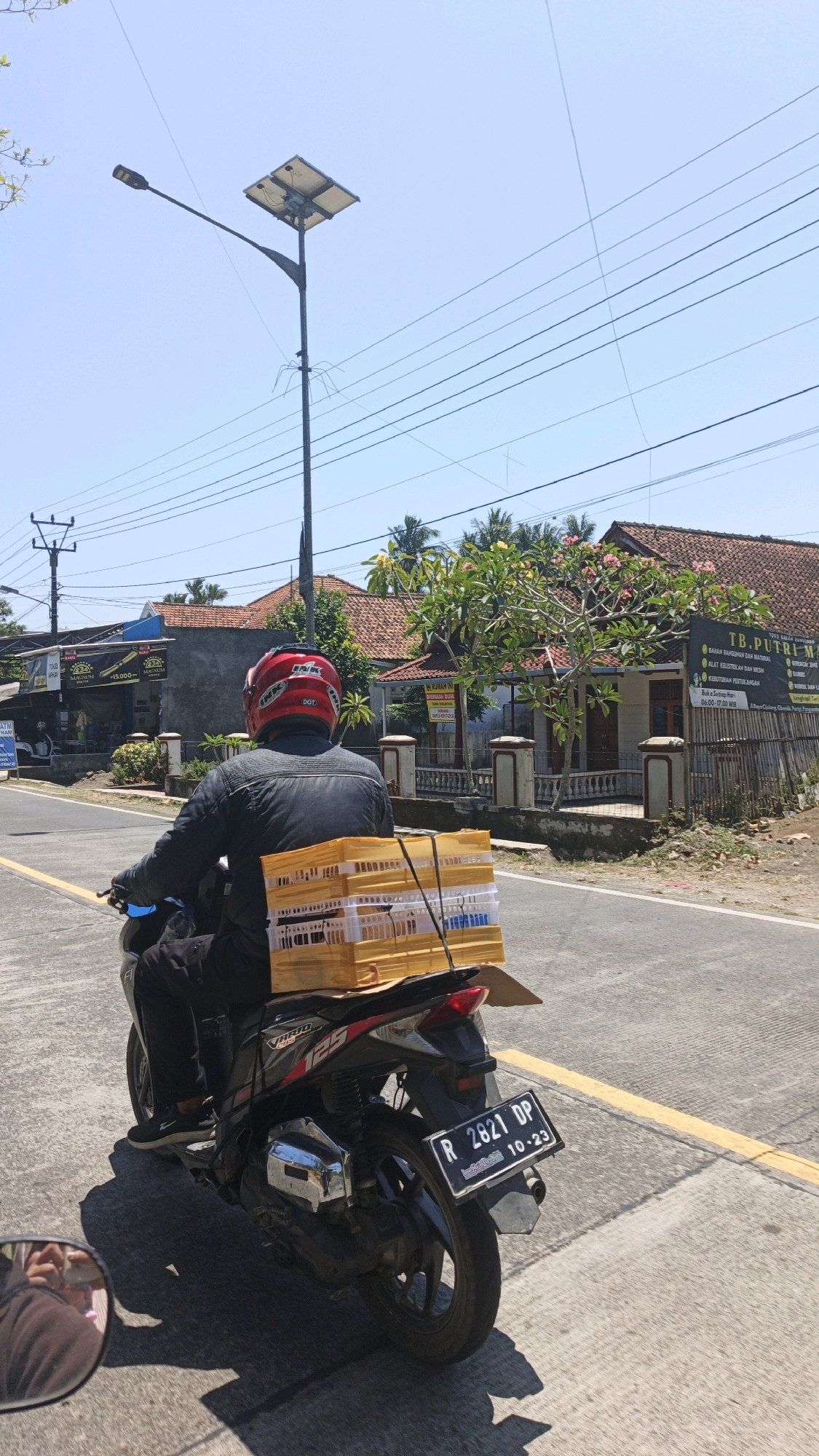 A person wearing a black jacket and red helmet is riding a black motorcycle with a yellow cargo basket on the back, traveling on a road in Cibenda, Indonesia. The cargo basket contains stacks of plastic crates, possibly for delivering goods. The motorcycle has a license plate that reads "R 2821 DP 10-23". The environment is a rural or semi-urban area with buildings, trees, and power lines visible. The sky is clear and blue, indicating a sunny day. The road appears to be asphalt with a yellow line marking the lane. There are reflections in the motorcycle's rearview mirror, showing a person holding a phone.