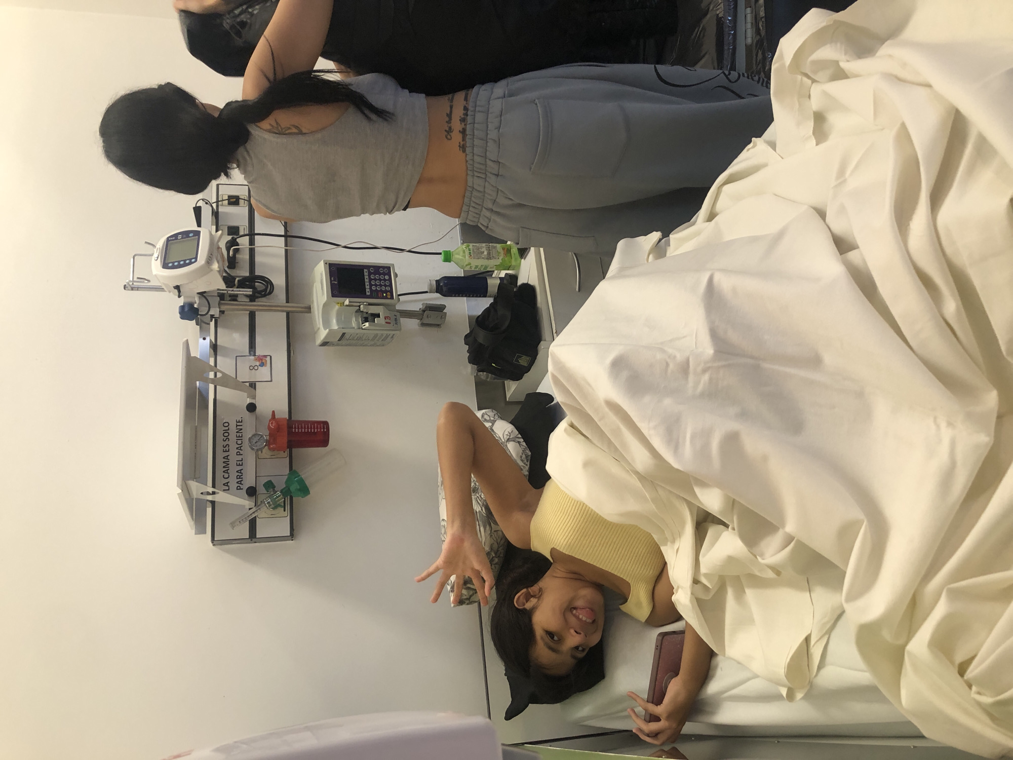 The image shows a person in a hospital bed covered in white sheets. Another person, wearing a gray tank top and gray sweatpants, is standing nearby, possibly tending to medical equipment. Medical equipment is mounted on a wall. A young girl is visible in the bed making a face. The scene appears to be indoors, possibly in a hospital room, in Buenos Aires, Argentina, since medical instructions are shown in Spanish.