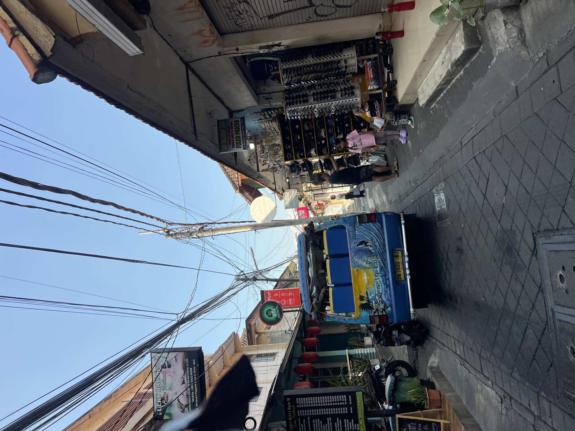 The image depicts a narrow street in Kuta, Indonesia, likely a tourist area, with buildings lining both sides. Overhead, a dense network of electrical wires is visible against a clear blue sky. On the left, a building with an orange-tiled roof has a sign for "LUCKY BALI Spa 9 Body Treatment" displaying prices and contact information. Further down the street on the left, red lanterns are hanging.
In the center of the street, a blue van, decorated with a surfing scene and labeled "party bus," is parked. Its rear license plate reads "DK 7033 BJ." Two motorcycles are parked on the left side of the street.
On the right side of the street, a shop displays shelves filled with sunglasses and hats. Two people, a man and a young girl wearing a pink shirt and purple sandals, are standing in front of the shop. Another person, partially obscured, is seated on a bench. Bottles of liquid are placed on the ground near the shop entrance. The street is paved with dark grey interlocking bricks, and the ground appears wet in some areas.